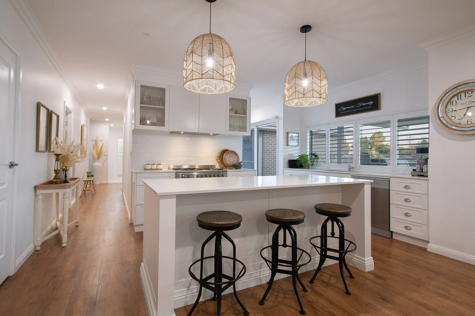 Modern white kitchen with two woven pendant lights, a white island with three wooden barstools, and a window with plantation shutters.