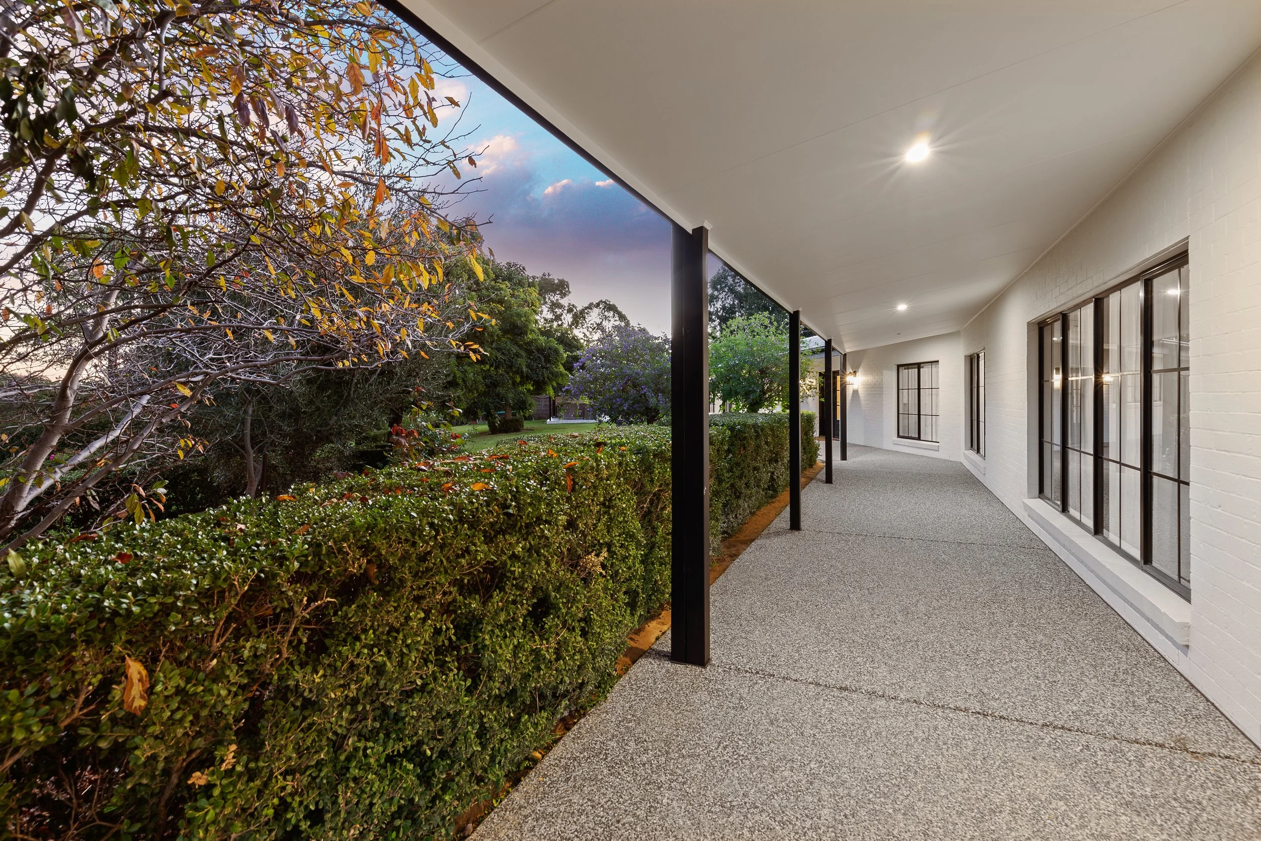 Exterior view of a white brick building with large windows, a covered walkway, and landscaped garden with shrubs and trees at dusk.