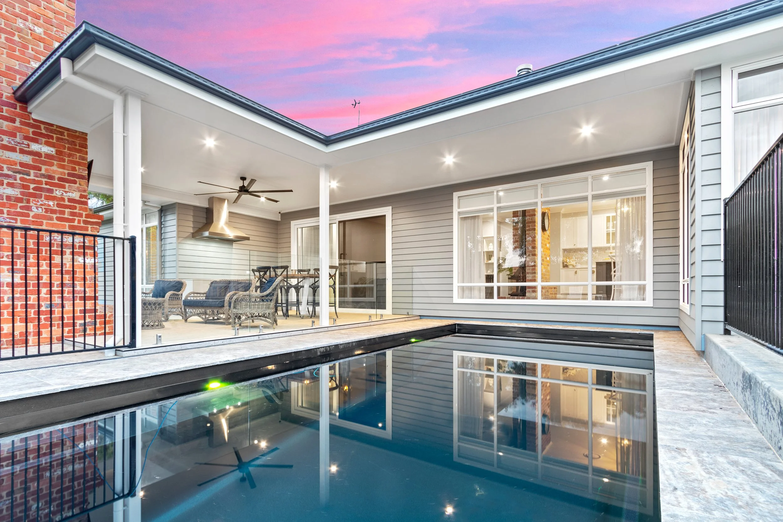 Modern backyard with a swimming pool, outdoor seating, covered patio, and a sunset sky.