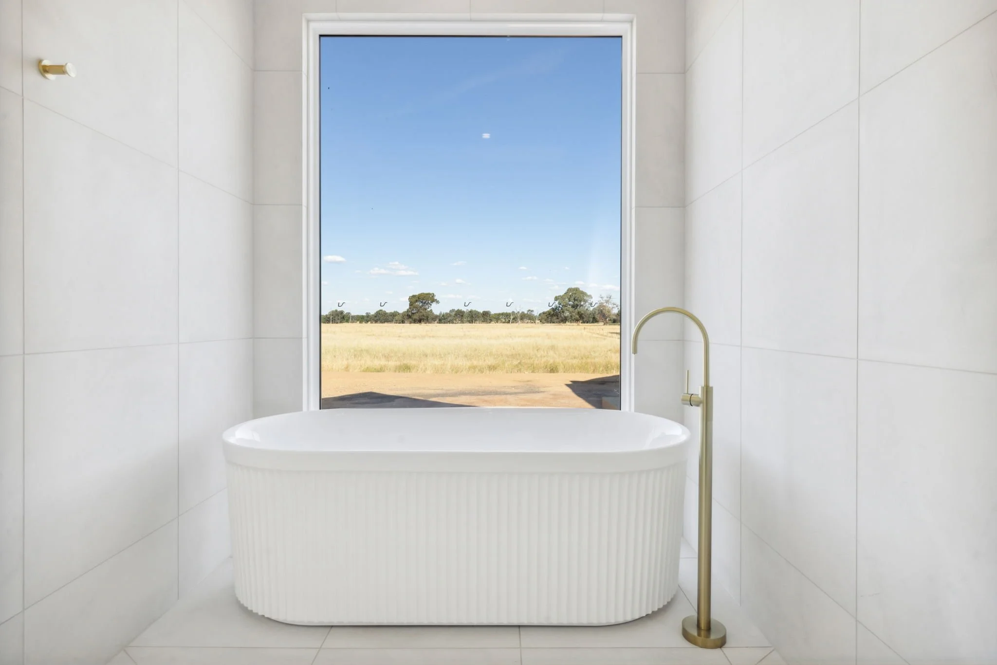 A modern bathroom with a white freestanding bathtub and a golden floor-mounted faucet, large window showing an outdoor landscape of a grassy field and trees under a blue sky.