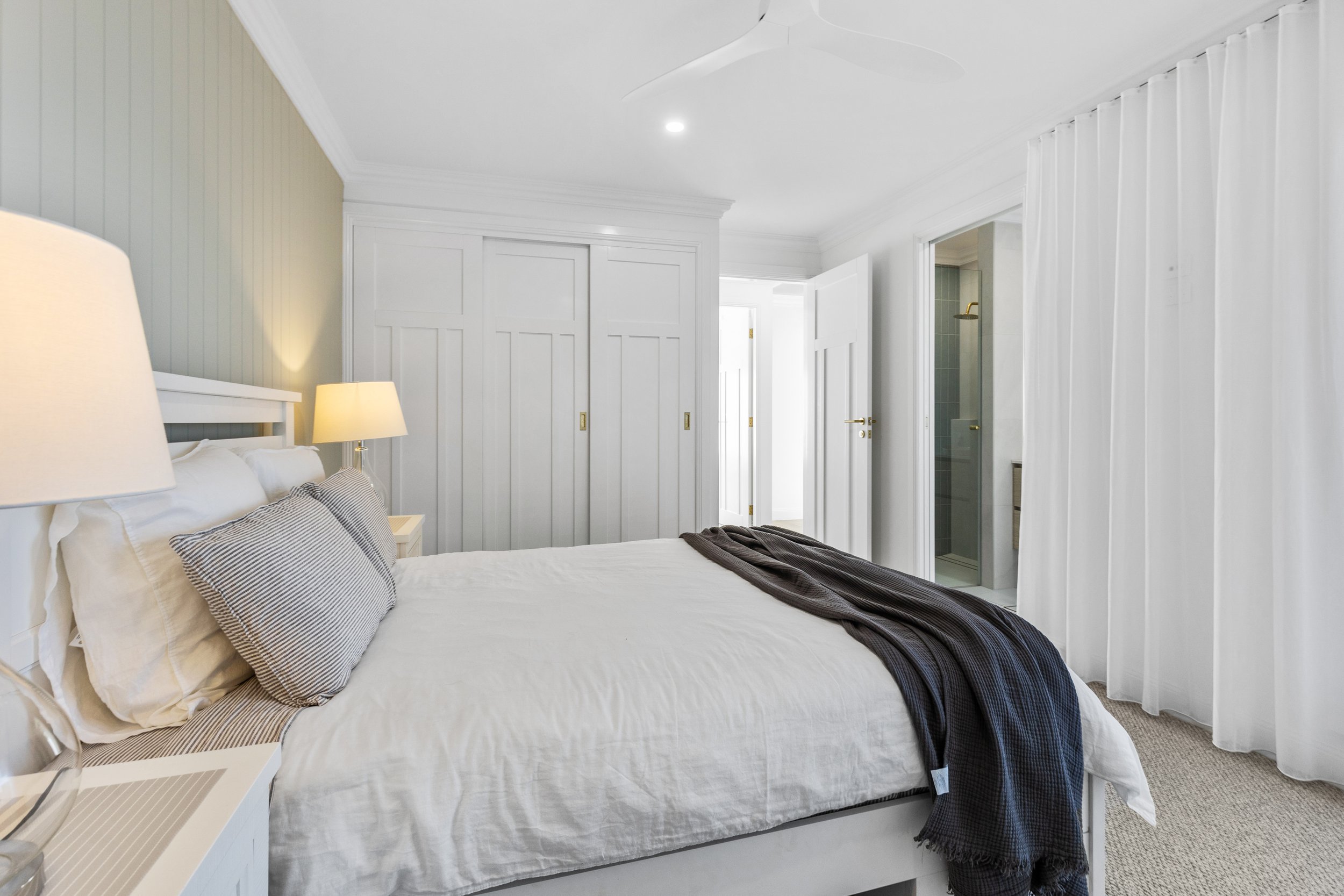 A bedroom with white paneled walls, a bed with white bedding and pillows, and sitting on a beige carpet, with nightstands and lamps, a closet, and an open door to a bathroom.