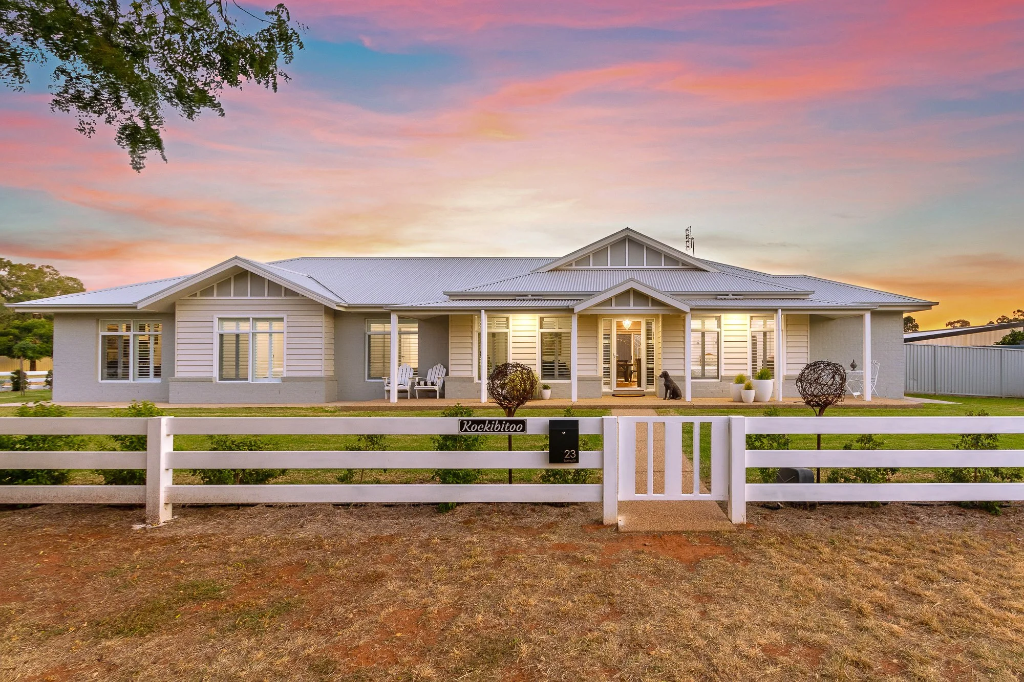 A modern single-story house with white siding and a light-colored roof, surrounded by a white fence with a gate. The house is set against a colorful sunset sky with pink, orange, and blue hues. There are outdoor seating areas with chairs and decorative plants on the front porch, and some small trees and bushes in the yard.
