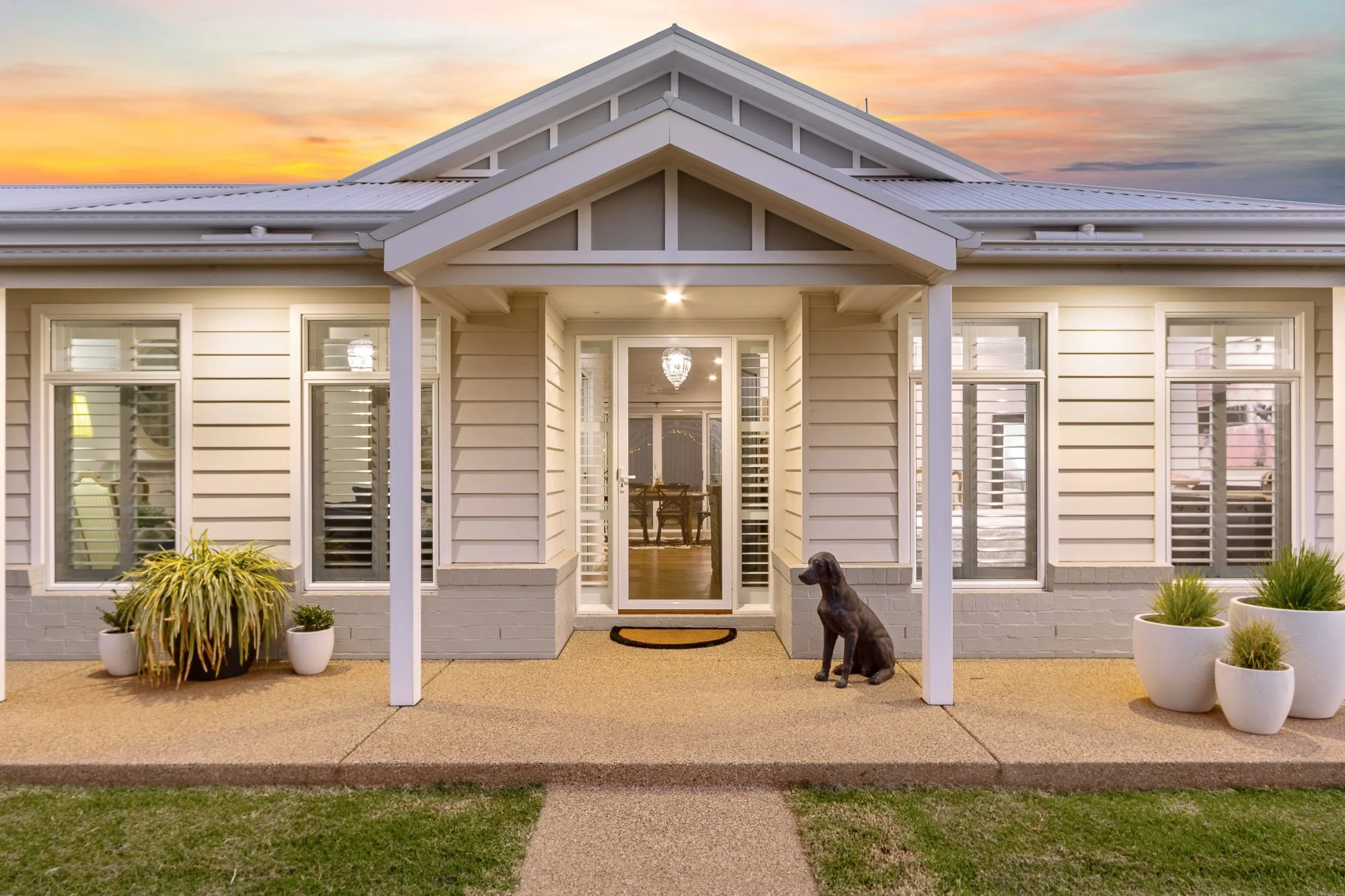 Front view of a house with cream siding and white trim, a small front porch, potted plants, and a dog statue, during sunset.
