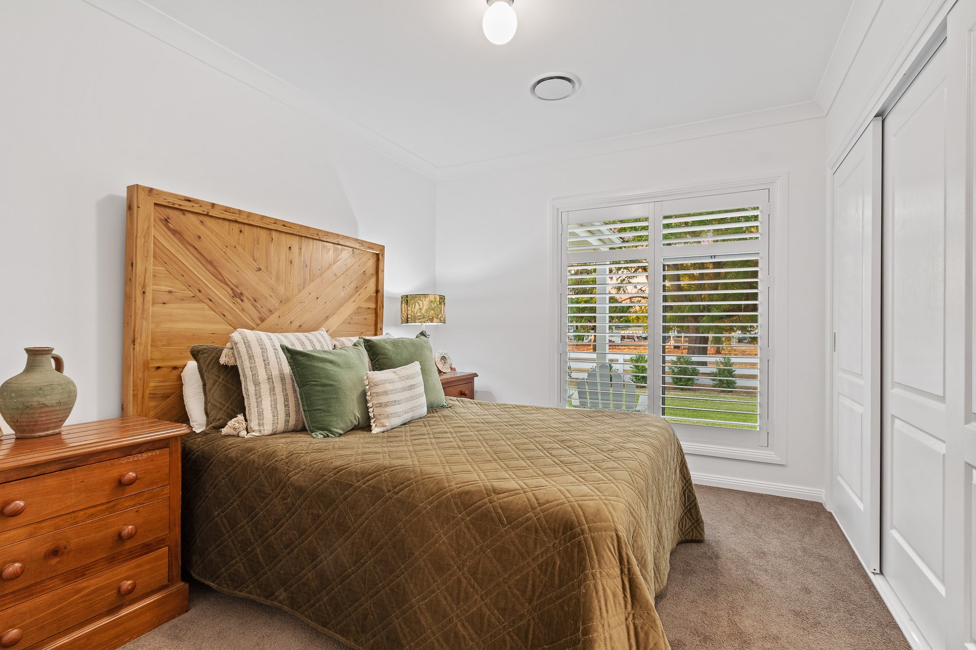Bedroom with a large wooden headboard, green and beige pillows, bedside table with a lamp, window with white blinds, white walls, and sliding closet doors.