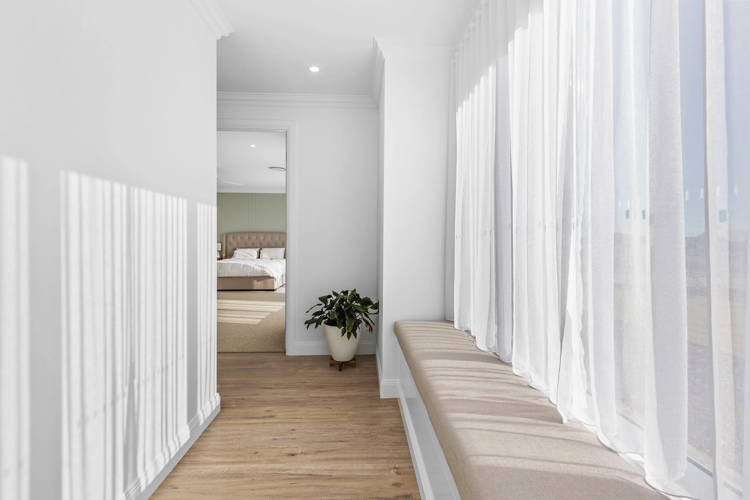 Bright hotel corridor with white walls, a built-in window seat with beige cushion, sheer white curtains, and a potted plant, leading to a bedroom with a bed and headboard visible in the background.
