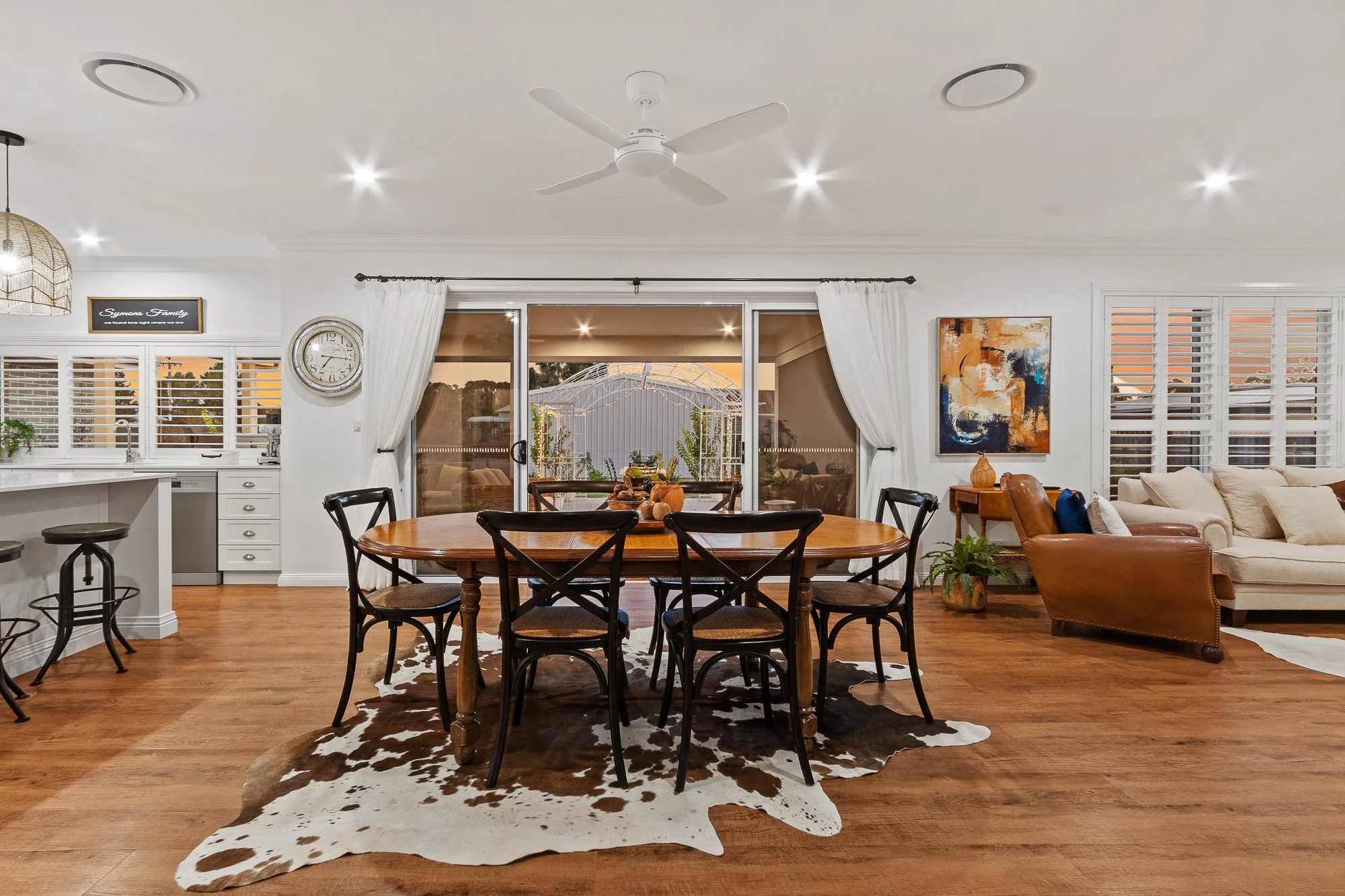 Open-concept living and dining room with wooden flooring, white walls, and large sliding glass doors leading to an outdoor patio, decorated with a round wooden dining table, black chairs, cowhide rug, beige sofa, leather armchair, artwork, and various potted plants.
