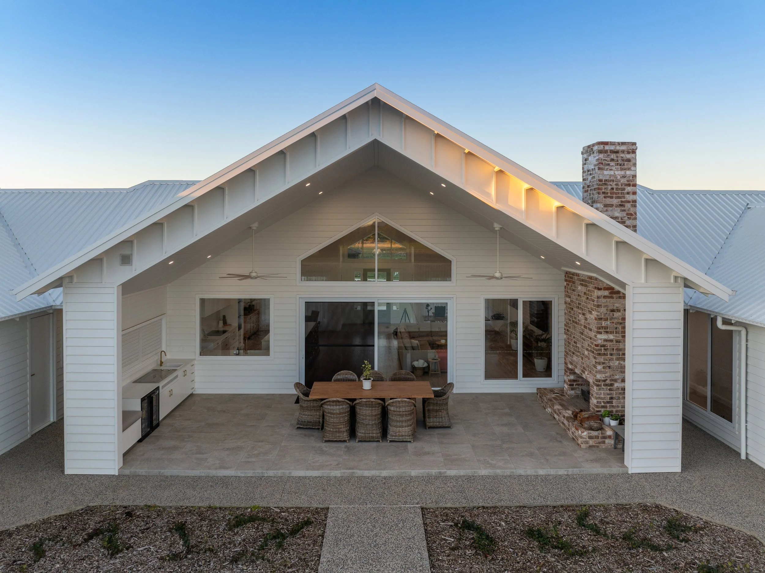 Backyard patio with outdoor dining table and wicker chairs, brick chimney, sliding glass doors, and ceiling fans, attached to a white house with a gabled roof.