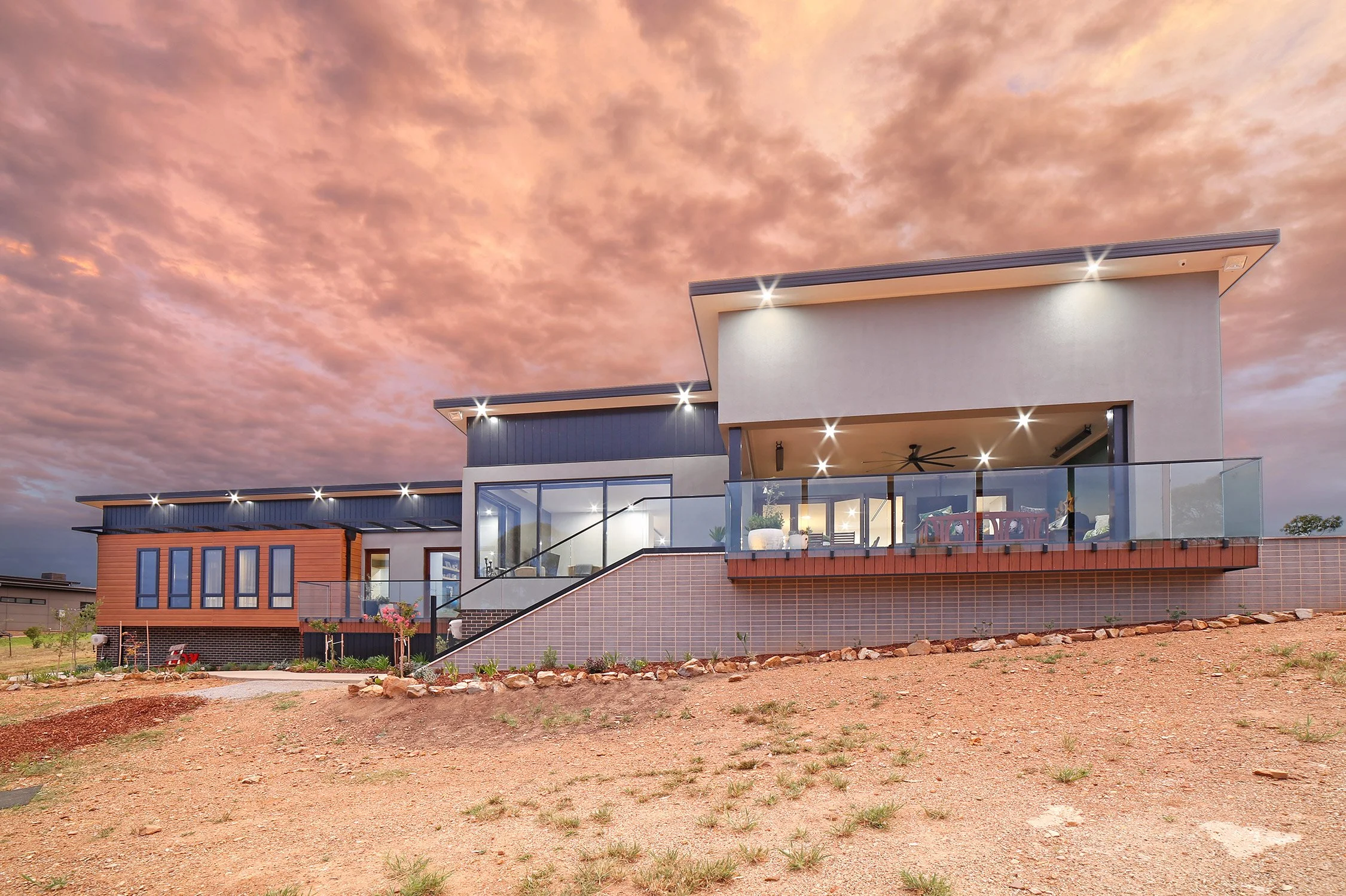 Modern house with multiple levels, large windows, and a spacious balcony, set against a dramatic pink and orange sunset sky.
