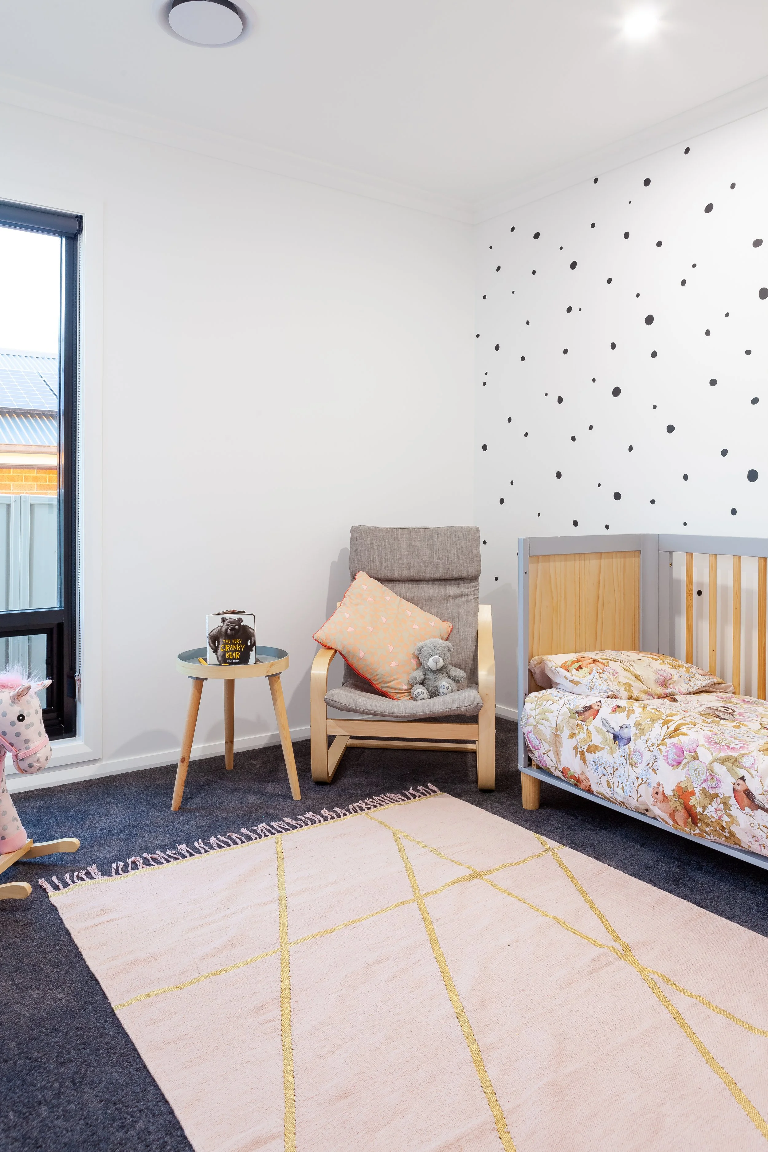A child's bedroom with a single bed, a beige rocking chair with a pillow and a teddy bear, a small round table with a book and a toy, a pink and white rug, a window, and a wall decorated with black polka dots.