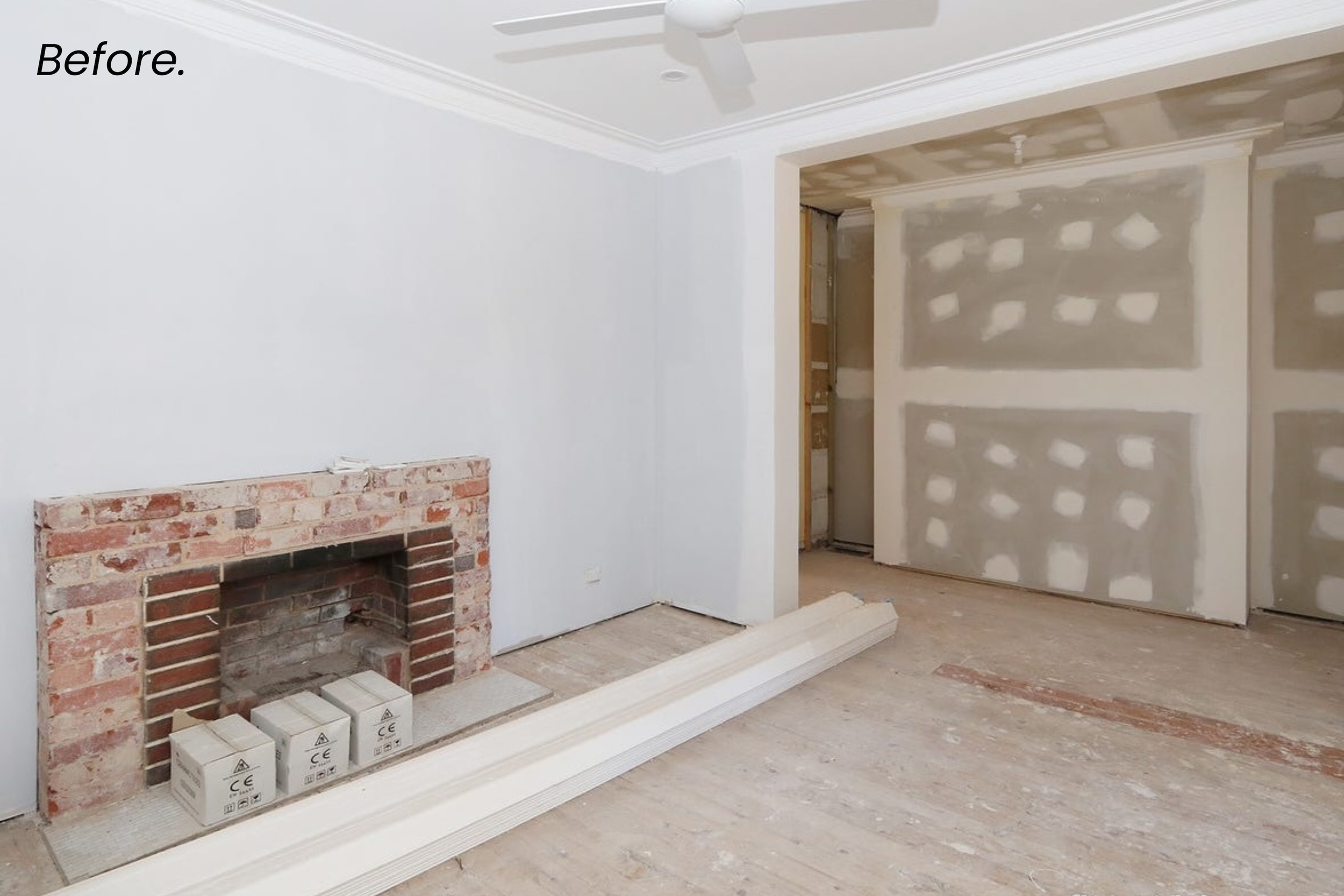 Empty room under renovation with white painted walls, unfinished drywall on one wall, and a brick fireplace with construction boxes in front. The ceiling has a fan and crown molding.