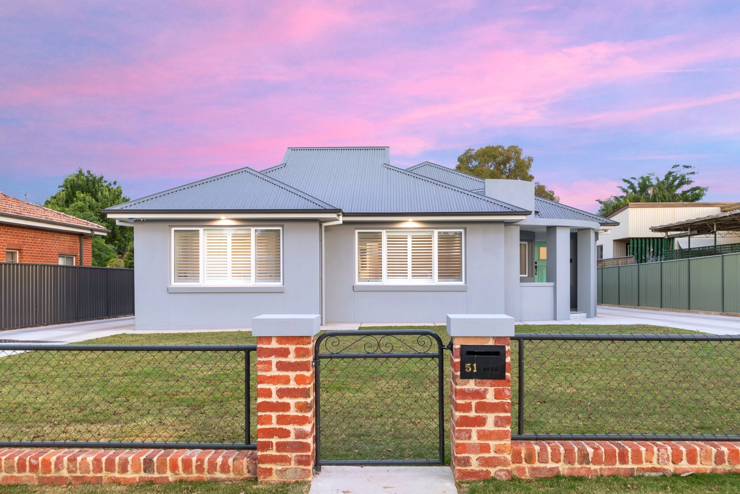 Front view of a modern single-story house with a metal roof, white exterior walls, and large windows with shutters, surrounded by a brick and metal fence, under a pink and purple sunset sky.
