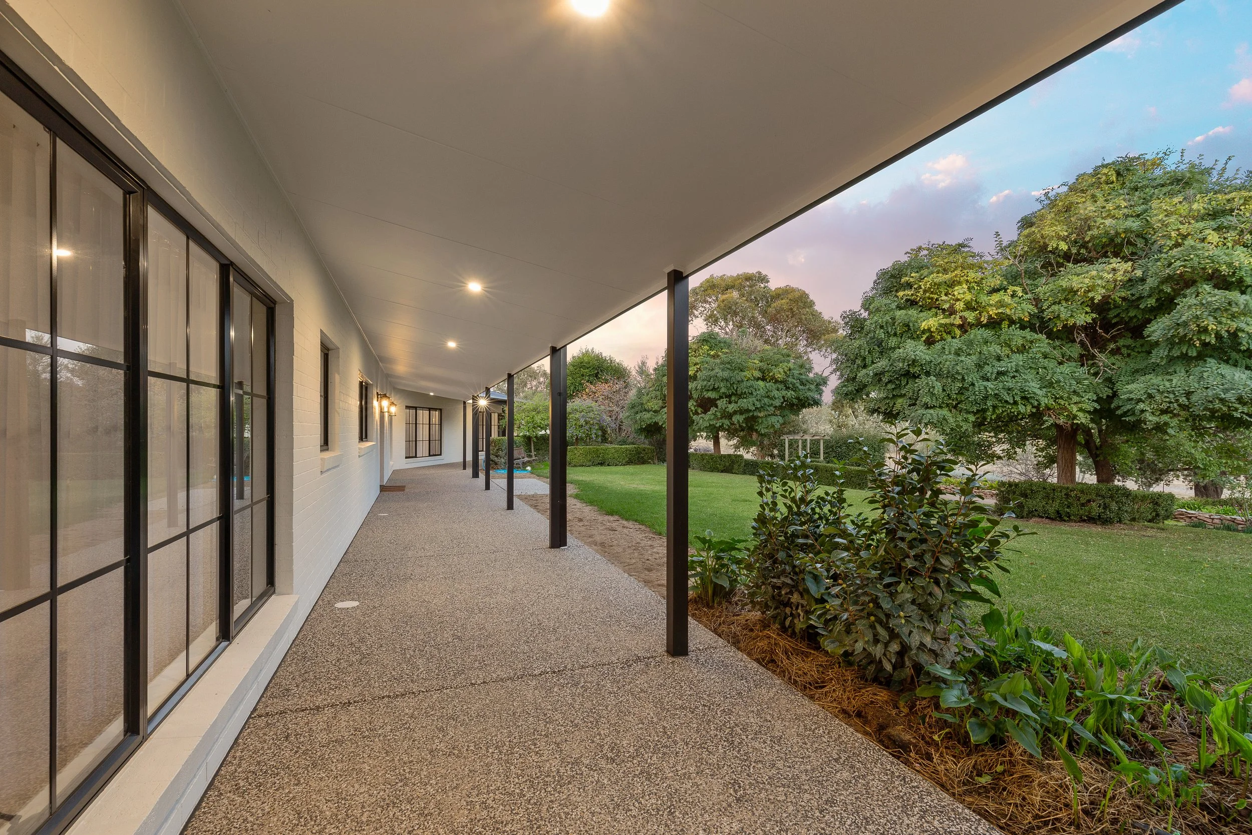 A covered porch with sliding glass doors on a white brick house, overlooking a landscaped yard with green grass, bushes, and trees at sunset.