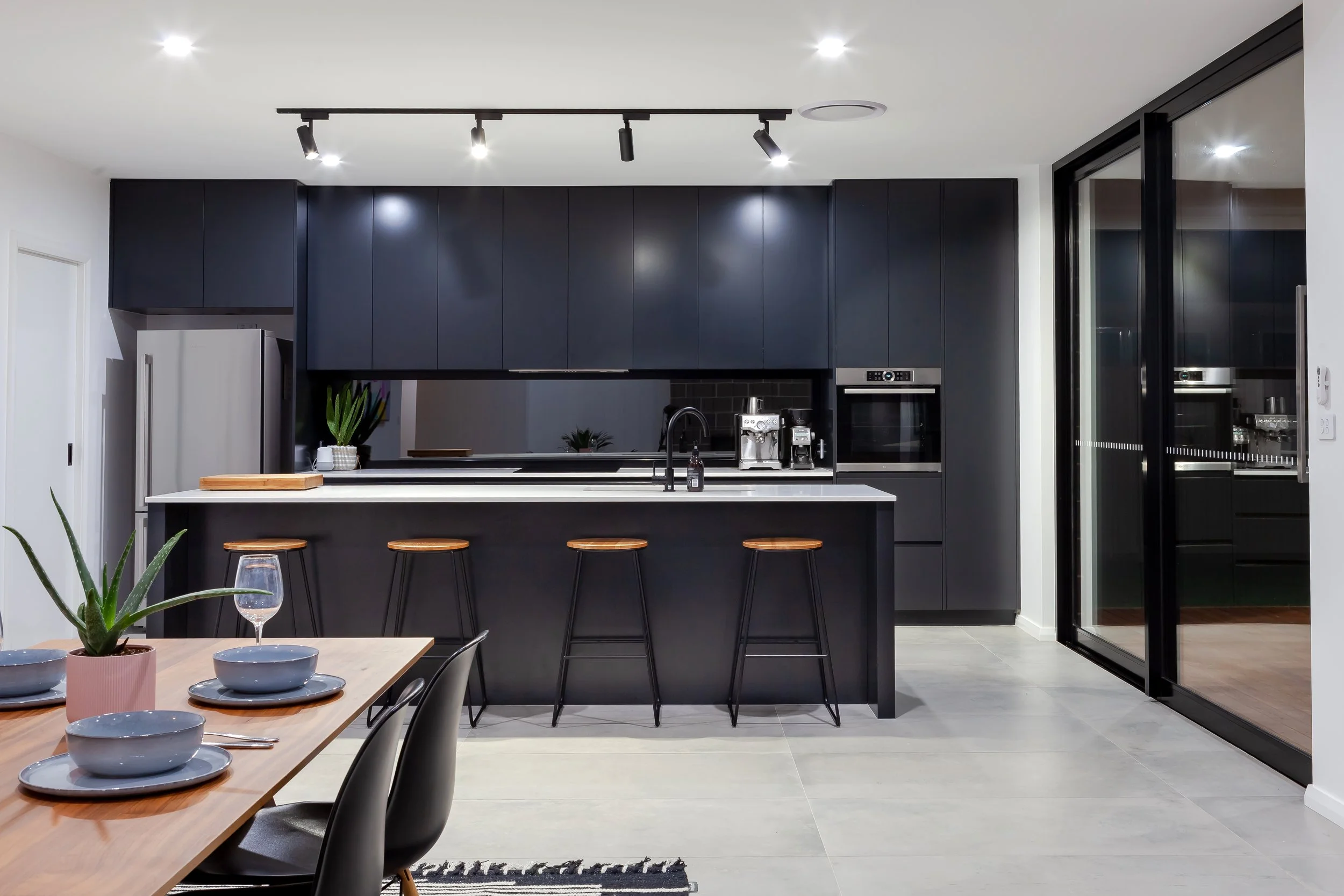 Modern kitchen with black cabinets, white countertops, and a kitchen island with three bar stools. There is a dining table set with bowls, plates, and glasses, and a glass sliding door leading outside.