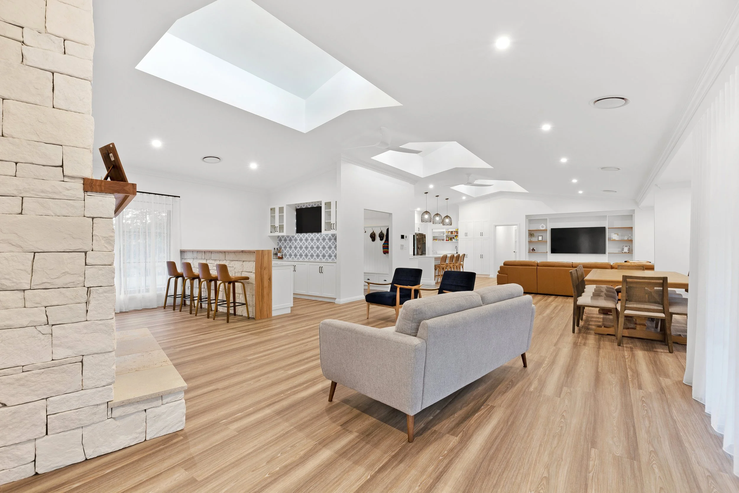 Spacious, modern living room with white walls, skylights, hardwood floors, and various seating options including a gray sofa, black armchairs, and a brown sectional sofa. Open-concept kitchen and dining area with white cabinetry, a large flat-screen TV, built-in shelves, and pendant lighting.