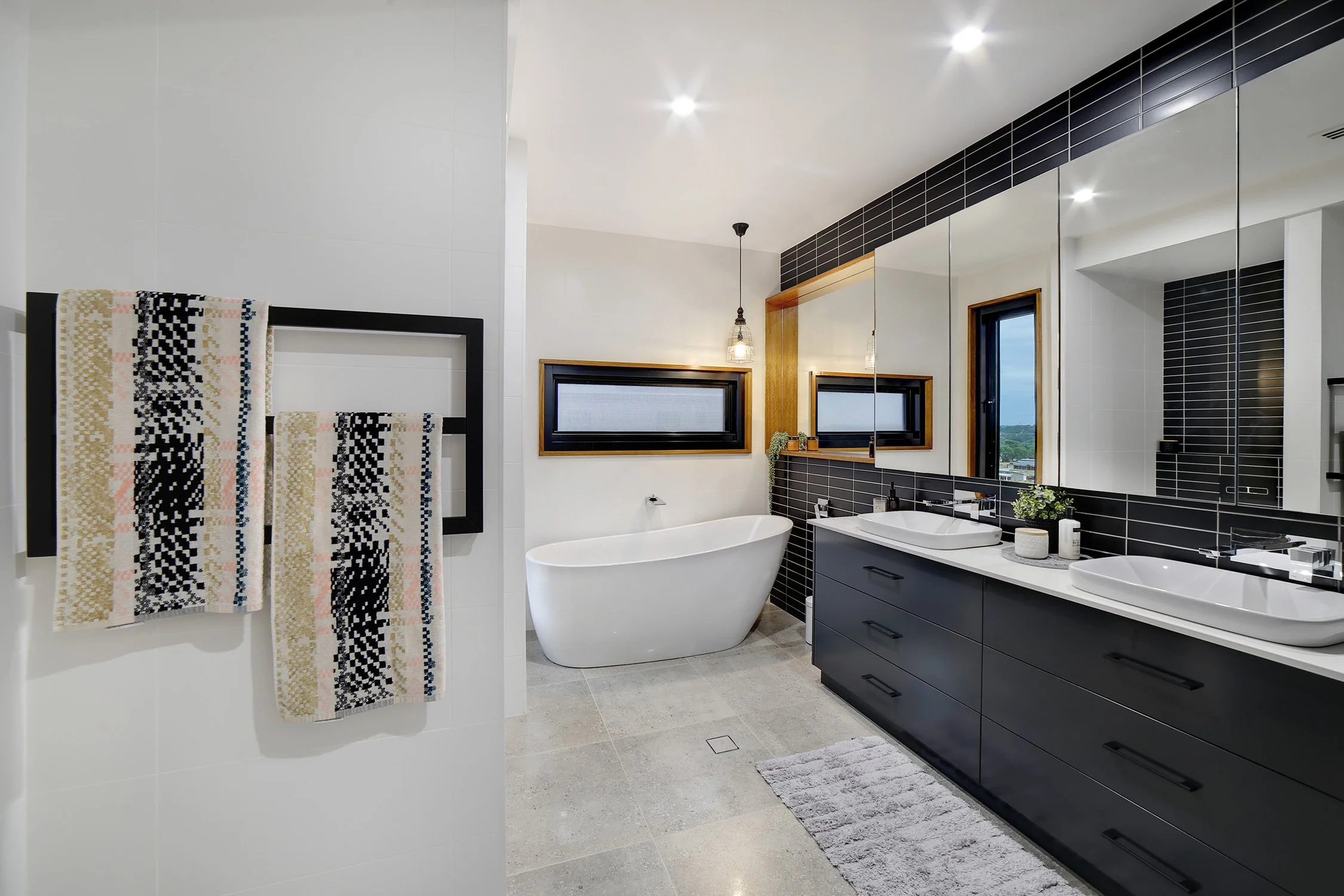 Modern bathroom with white freestanding bathtub, black vanity with dual sinks, large mirror, window, and towels hanging on a black towel rack.