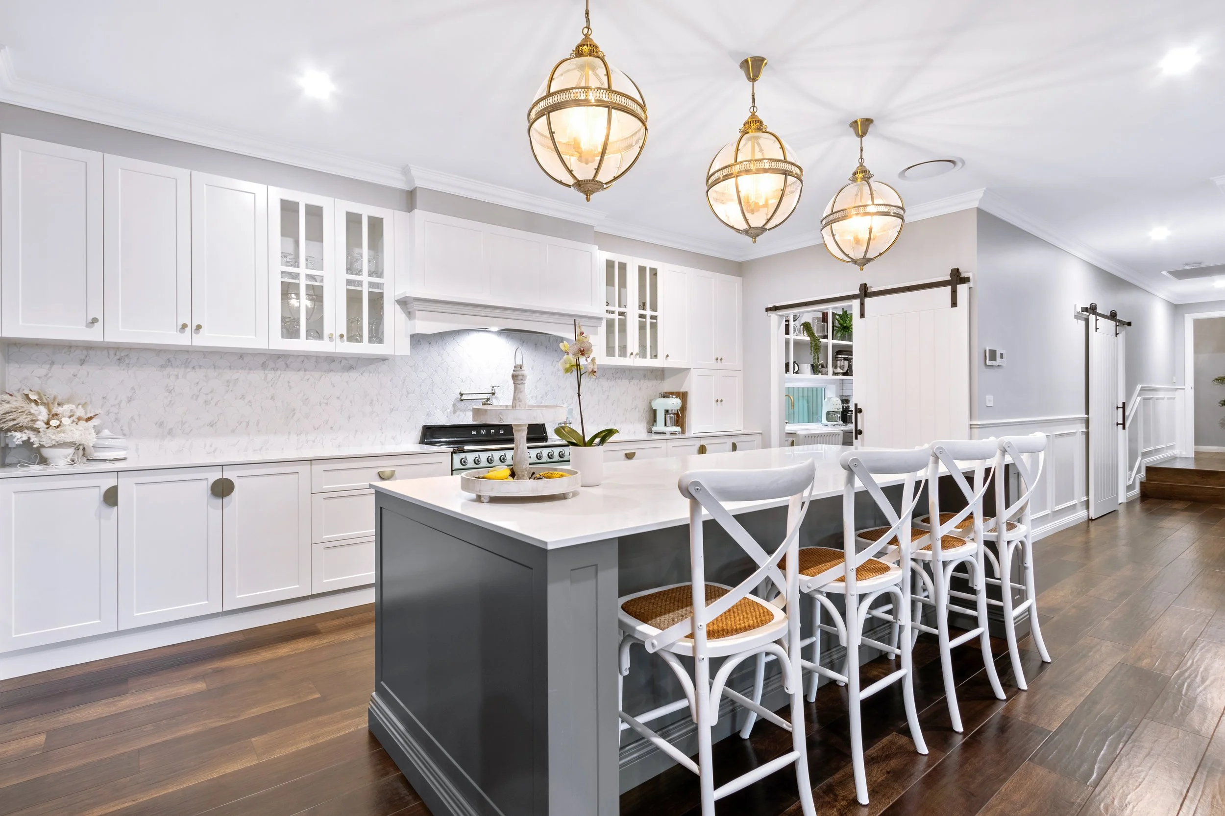 Modern kitchen with white cabinetry, a large central island with dark gray base, white countertop, and five white chairs with woven seats. Three pendant lights hang from the ceiling, and there is a sliding barn door to a pantry area. The flooring is dark wood, and the walls are light gray.