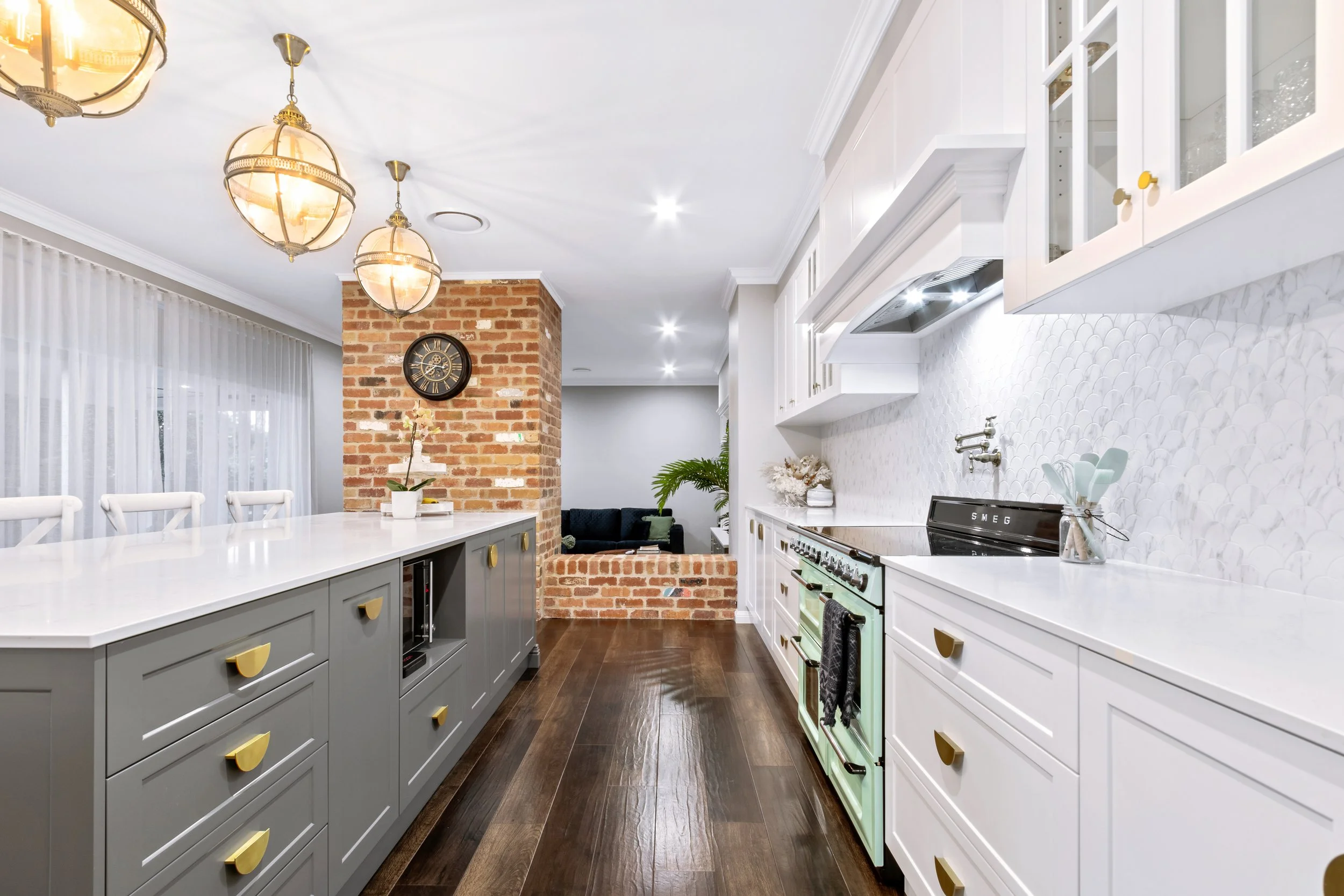 Modern kitchen with white cabinets, a green stove, and a long gray island with white chairs, brick wall, and hanging light fixtures.