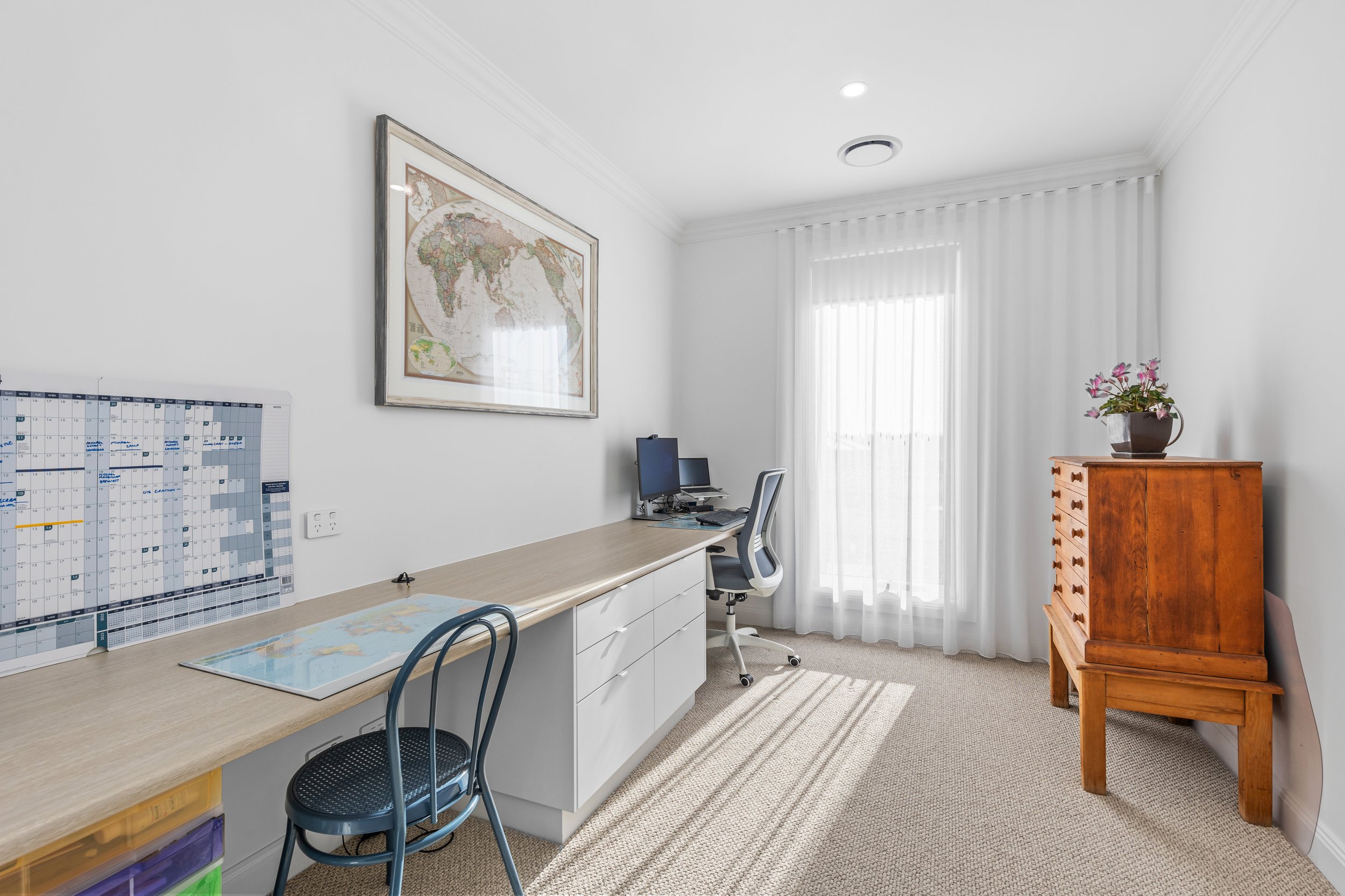 A bright, minimalist home office with a white wall, a long desk, a chair, a world map on the wall, a computer, a potted plant on a wooden cabinet, and sheer white curtains.