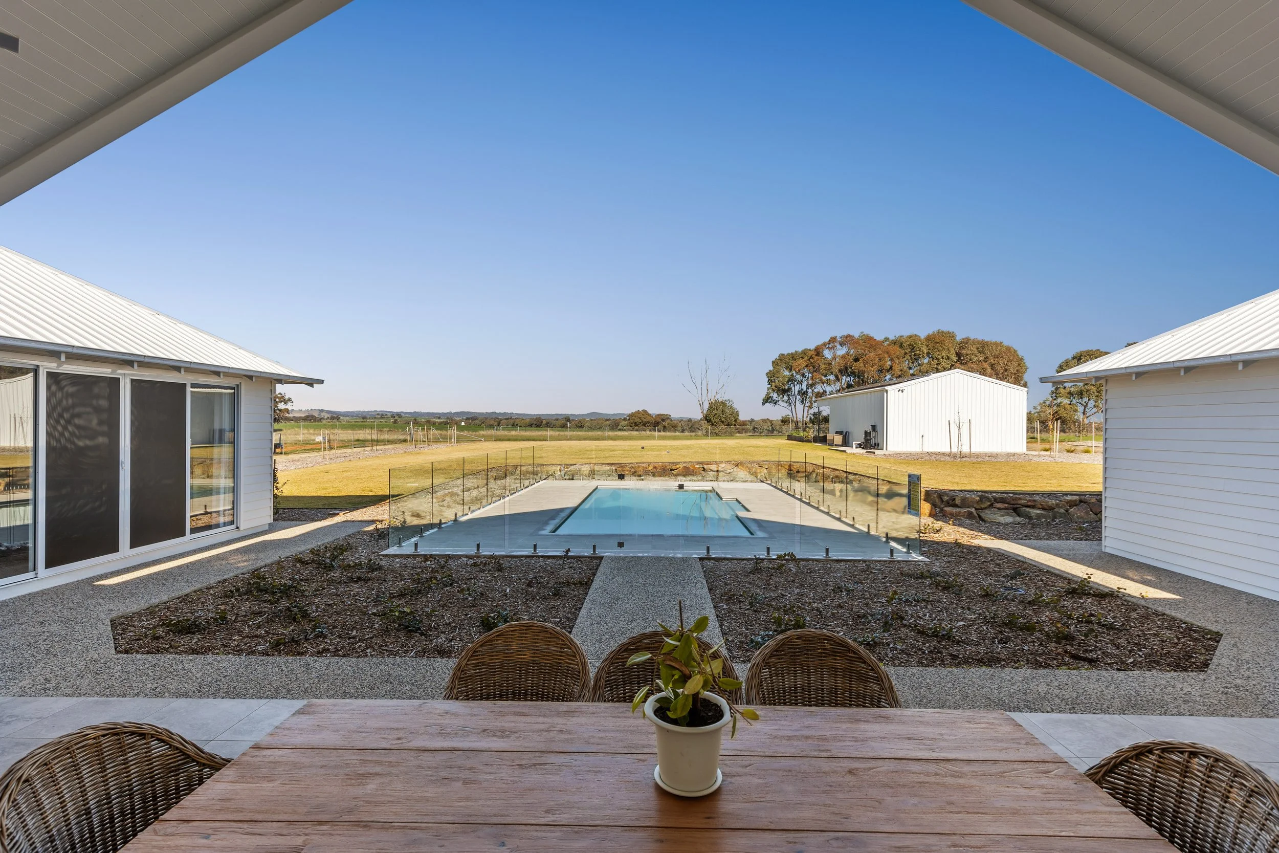 View through a covered patio towards a backyard with a swimming pool surrounded by glass fencing, two white houses with metal roofs on either side, and a landscape with trees and open fields in the distance.