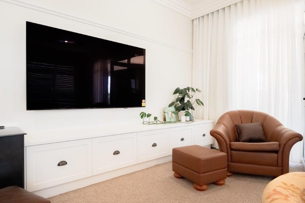 Living room with a large flat-screen TV mounted on the wall, a white built-in cabinet below, a brown armchair with a matching ottoman, a gray pillow on the armchair, a side table with potted plants, and white curtains covering a large window.