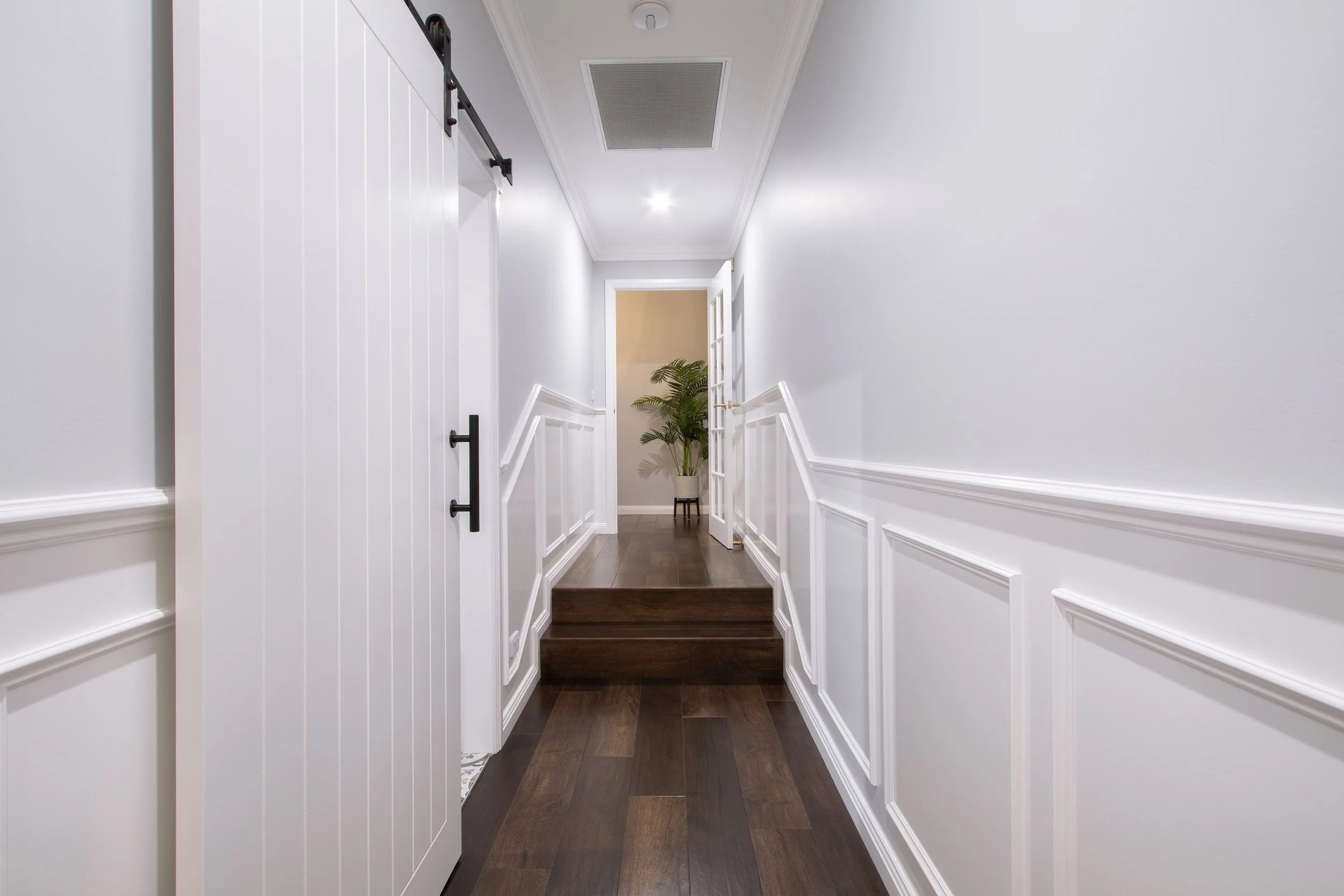 Interior hallway with white walls, dark wooden floors, a sliding barn door on the left, and a doorway at the end leading to a room with a potted plant.