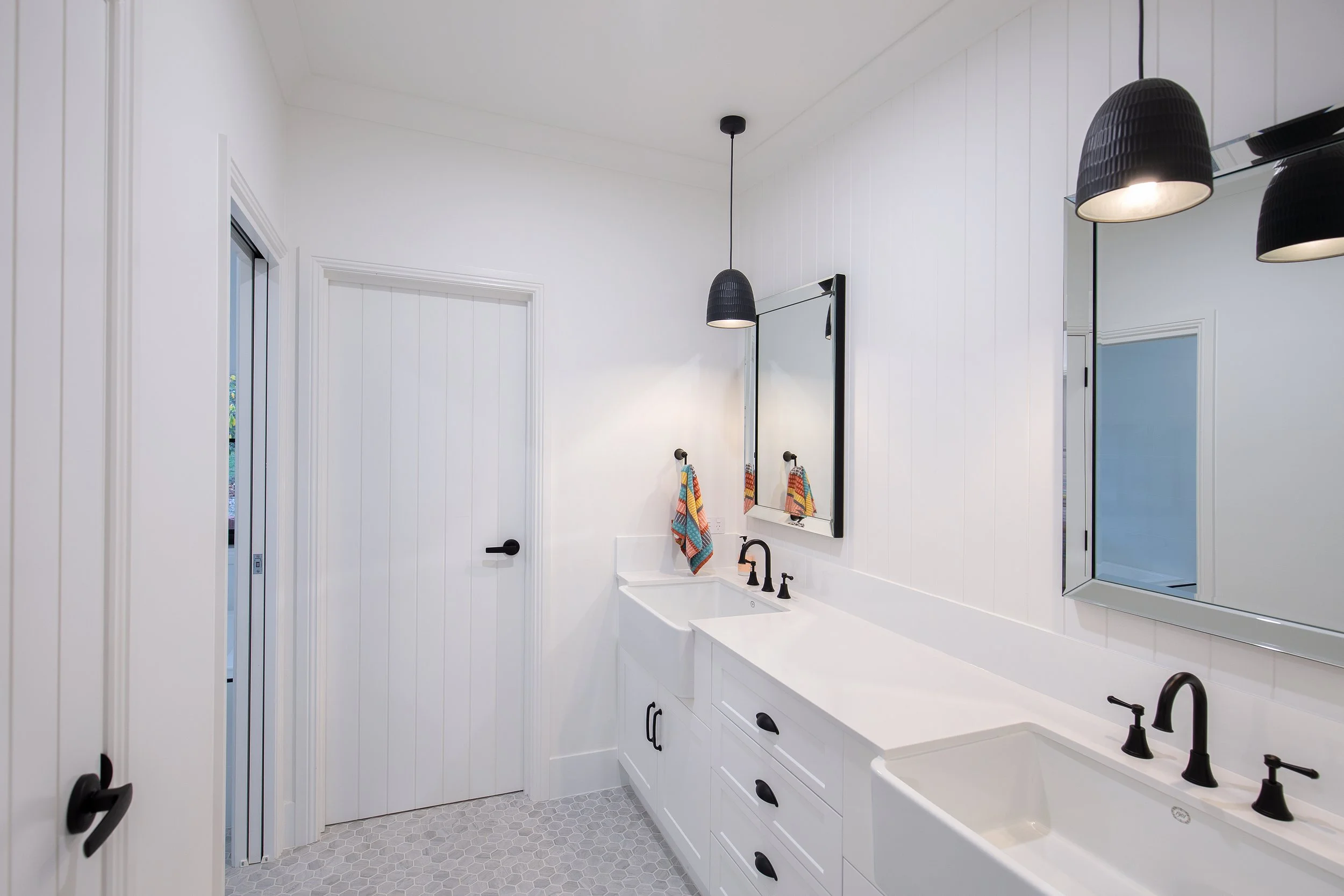 Modern white bathroom with black fixtures and twin sinks, black pendant lights, large mirror, white wall paneling, patterned tile floor, towel hooks with colorful towels.