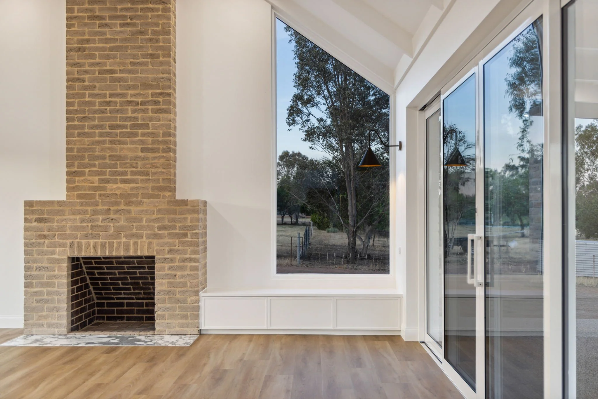 Interior of a room featuring a brick fireplace, large window, and sliding glass door leading outdoors, with hardwood flooring.