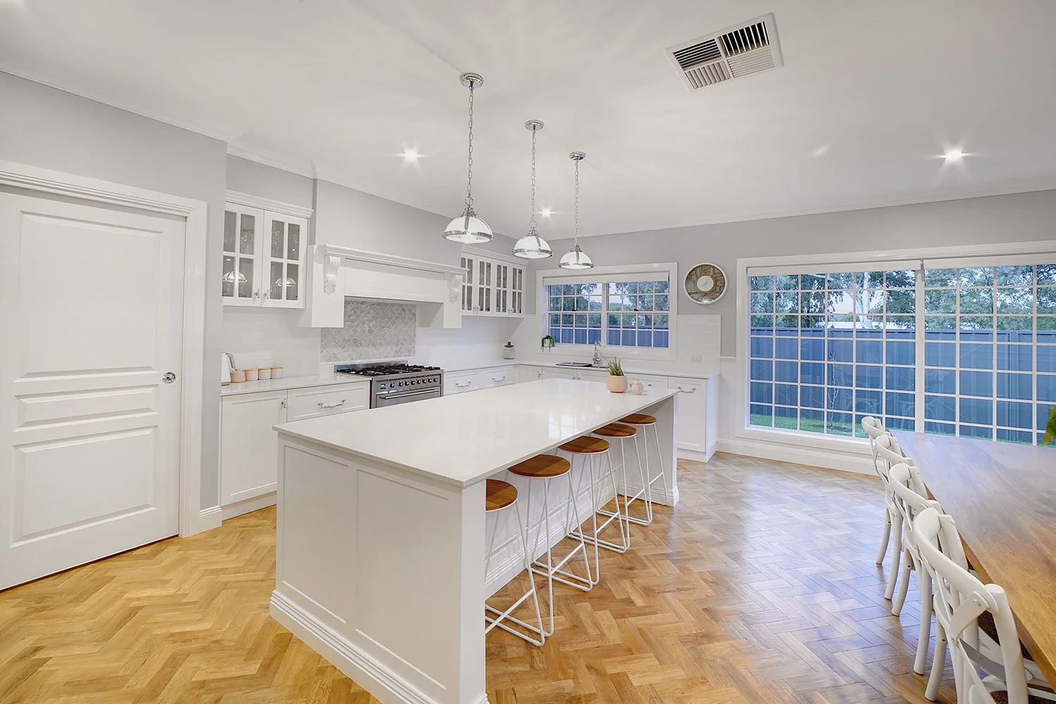 Bright, modern kitchen with white cabinetry, a kitchen island with wooden stools, stainless steel stove, large windows, and hardwood floors.
