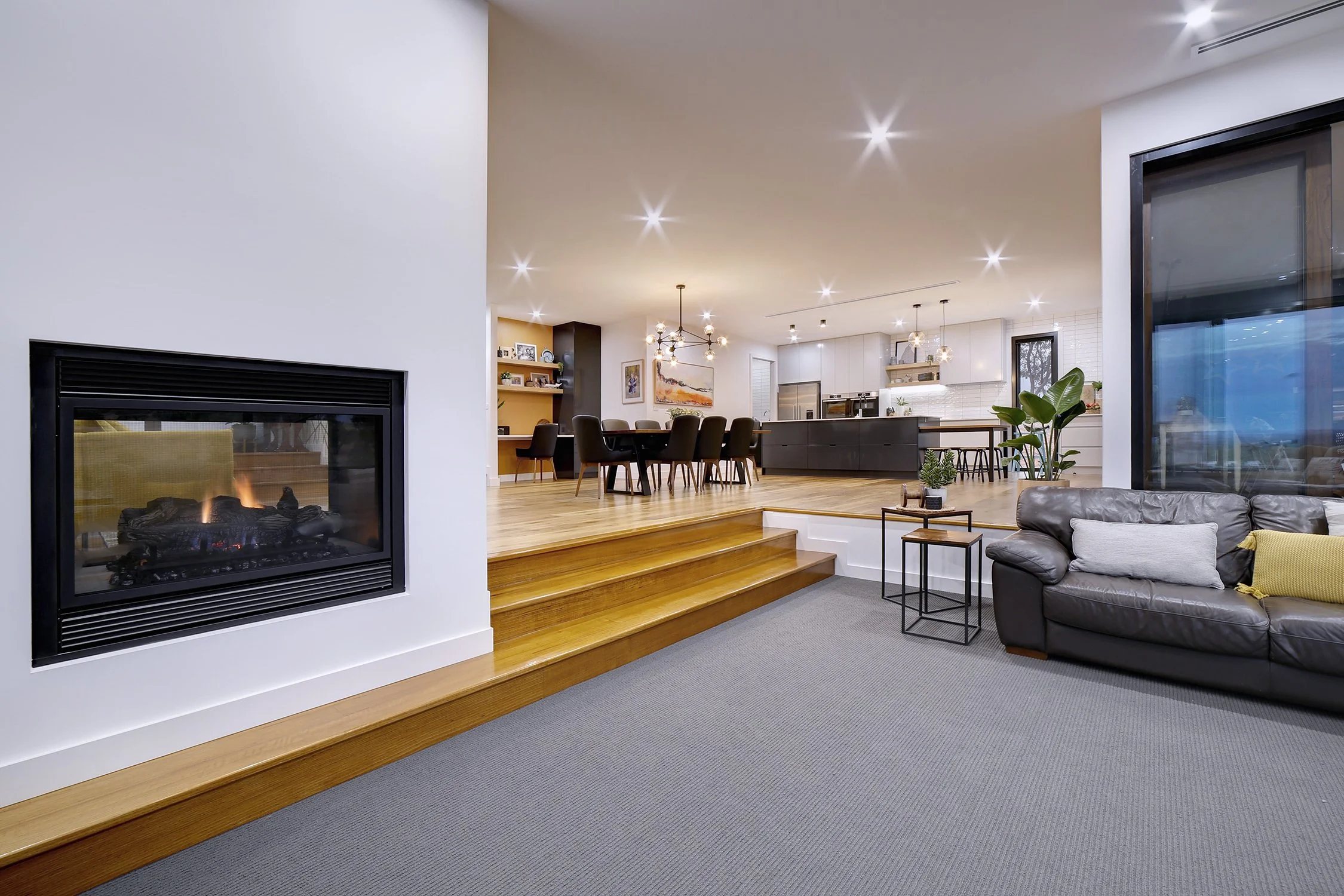 Bright living room with a gas fireplace, gray sofa with pillows, a small side table, and a large window. Elevated dining area with a black dining table and chairs, open kitchen with white cabinets, and pendant lights.