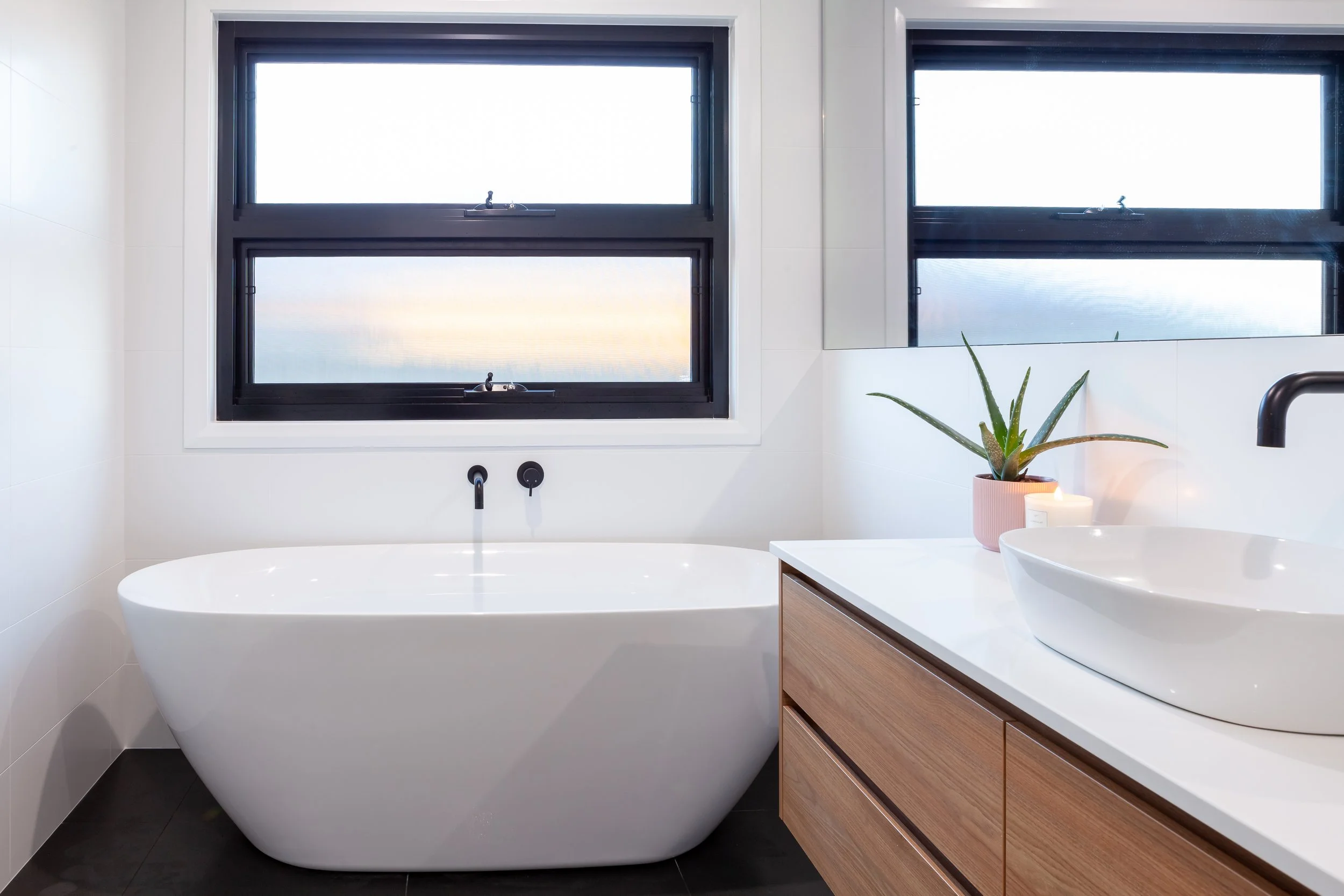 Modern bathroom with a white freestanding bathtub, two black-framed windows, a wooden vanity with a vessel sink, a potted aloe vera plant, a lit candle, and a soap dispenser.