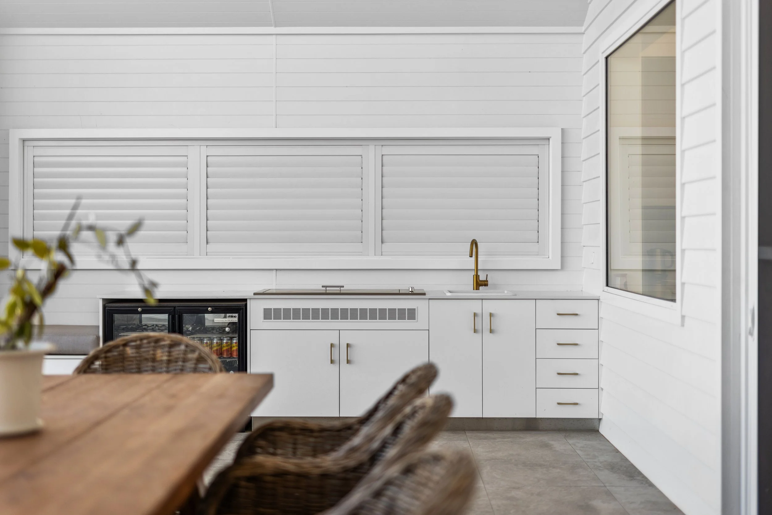Modern outdoor kitchen with white cabinets, a small black refrigerator, a window, and a wooden table with chairs in the foreground.