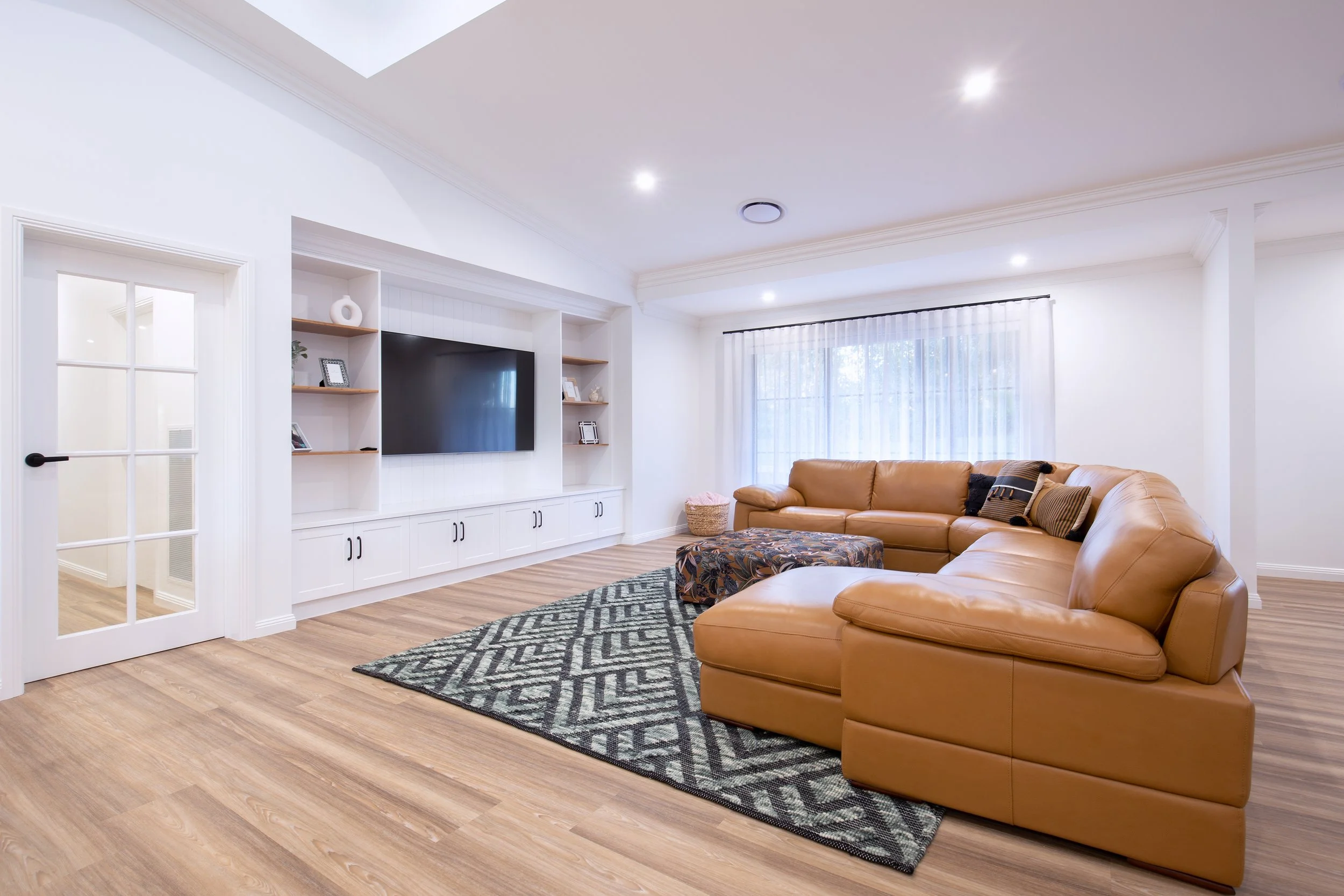 Living room with a large beige leather sectional sofa, a patterned area rug, a wall-mounted flat-screen TV, wooden shelves with decorative items, a large window with sheer white curtains, and light-colored wooden flooring.