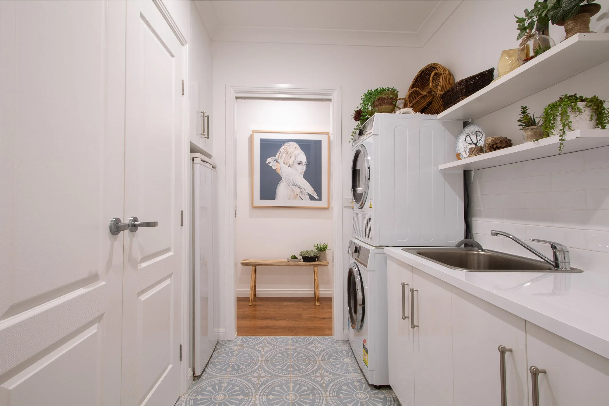 Laundry room with stacked washer and dryer, white cabinetry, open shelving with potted plants and woven baskets, sink, and a framed portrait of a woman with a bird on her shoulder hanging on the wall, and a wooden table with plants in the background.