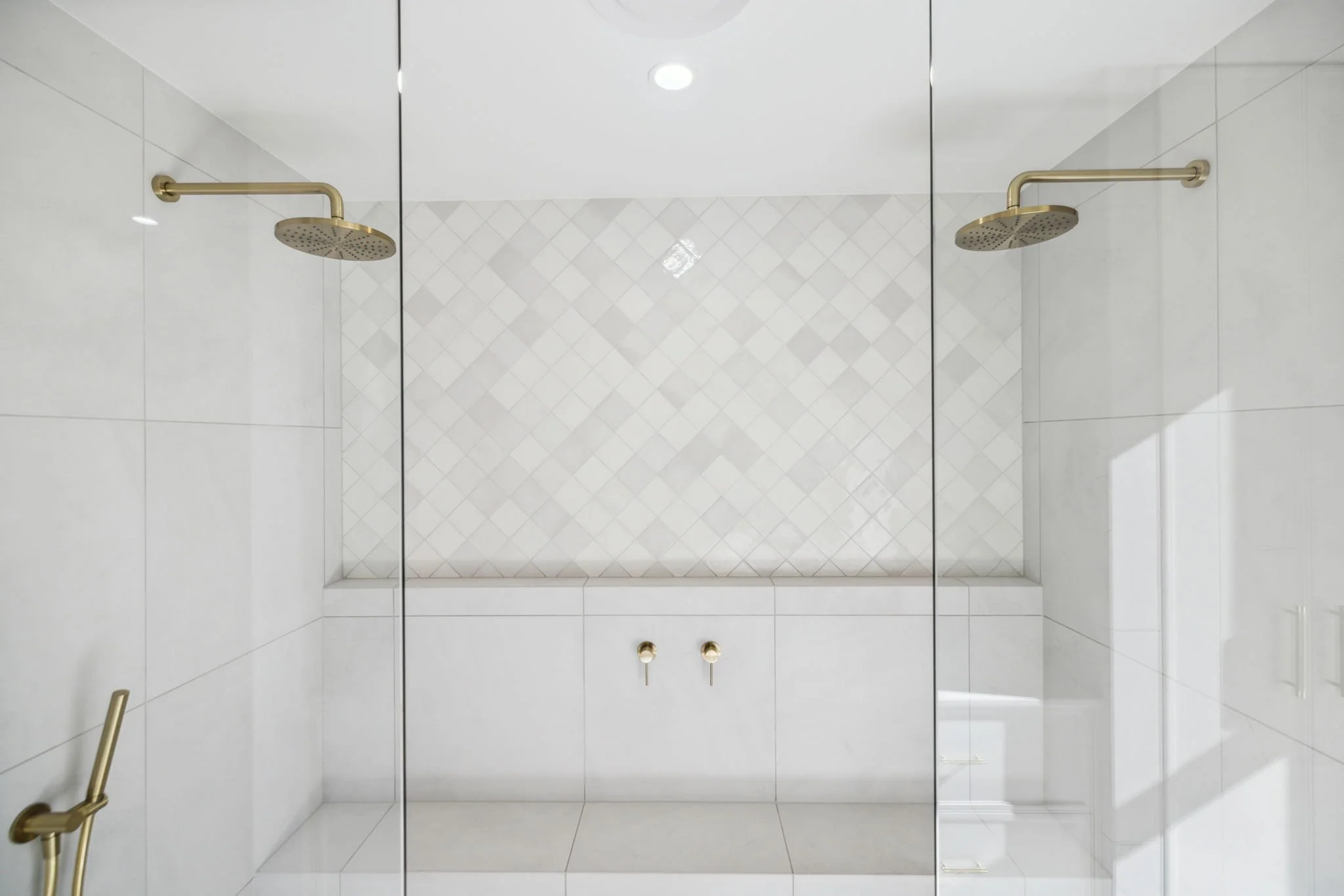 A modern walk-in shower with white tiles on the walls and a built-in bench. It has two gold-colored rain showerheads on opposite sides and a handheld showerhead with a gold handle on the left side. The shower is enclosed with glass panels.