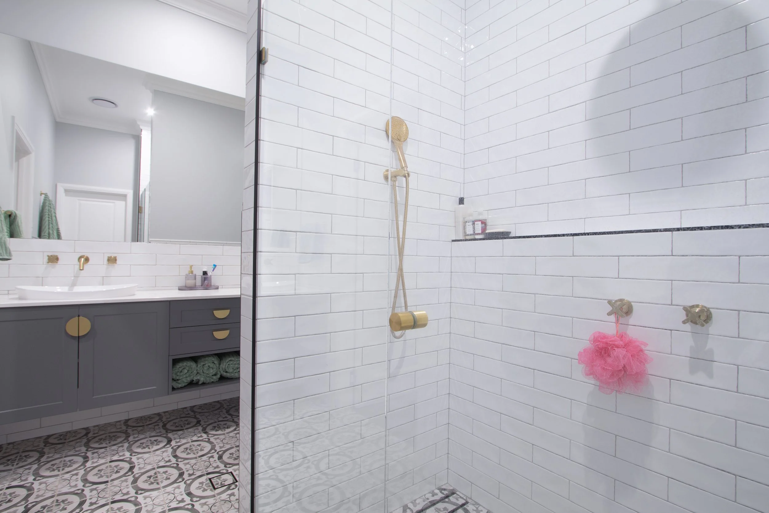 Modern bathroom with white tiled shower area, a showerhead, soap, and a pink bath pouf, with a vanity mirror and sink in the background.