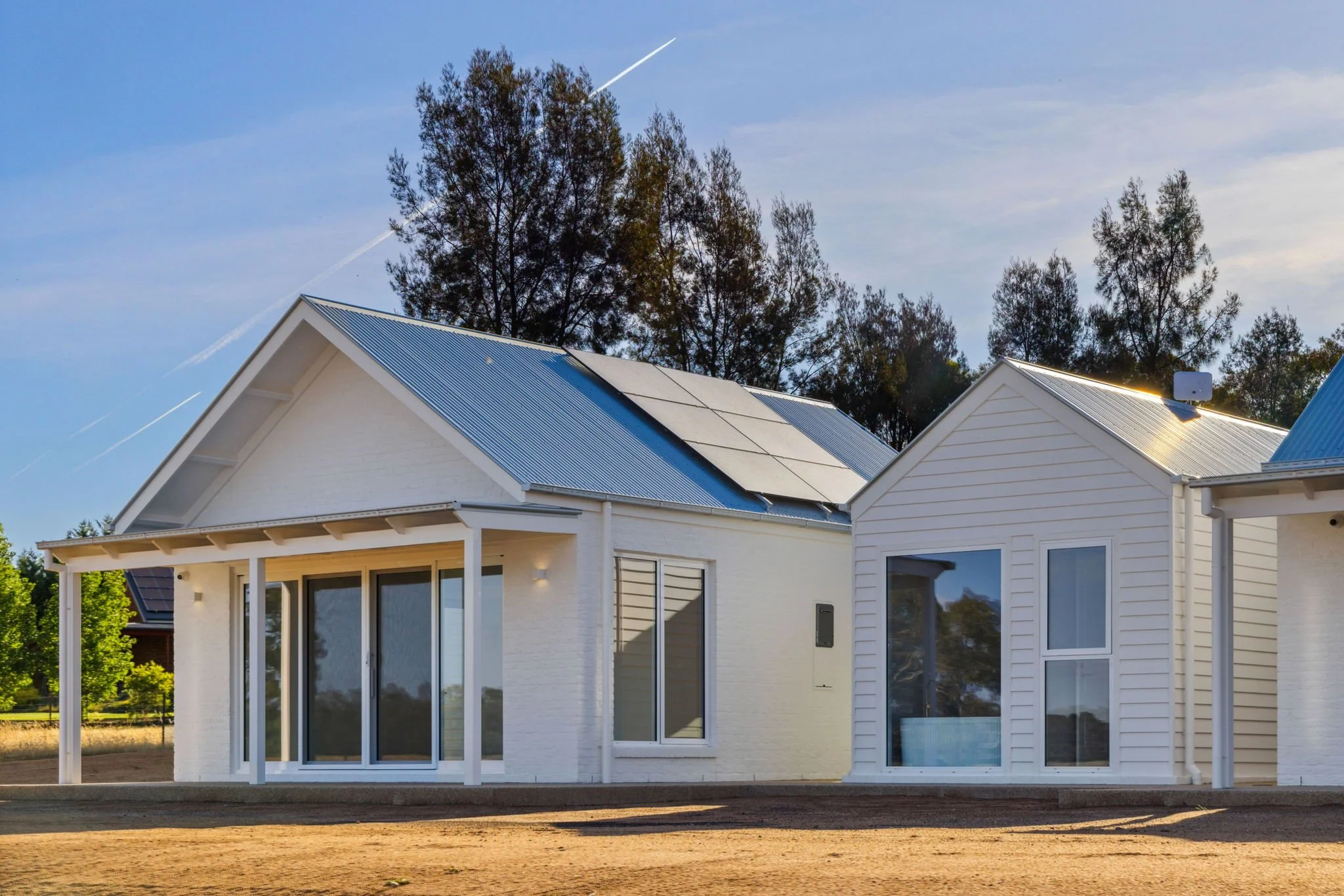 Modern white house with solar panels on the roof, large glass sliding doors, and a small porch, surrounded by trees under a clear sky.