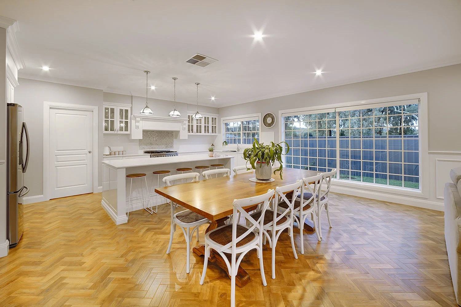 Bright dining area with a wooden table, white chairs, a potted plant, large windows with a blue fence outside, and a white kitchen in the background.