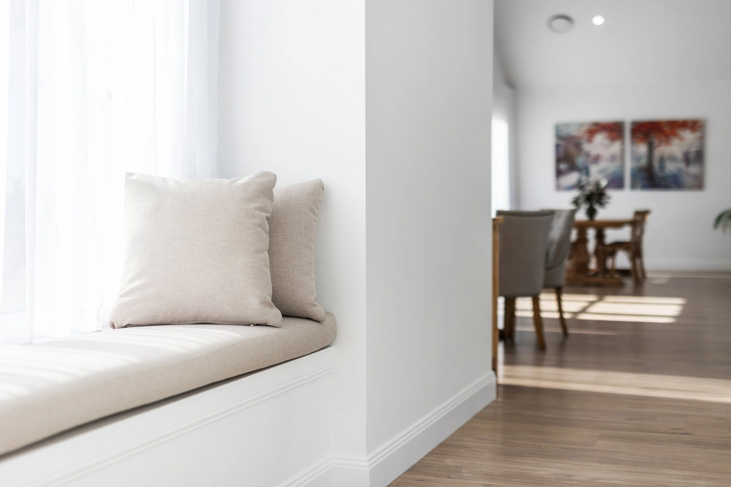 A bright living room with a built-in window seat with beige cushions next to sheer white curtains, and a dining area with artwork and a vase of flowers in the background.