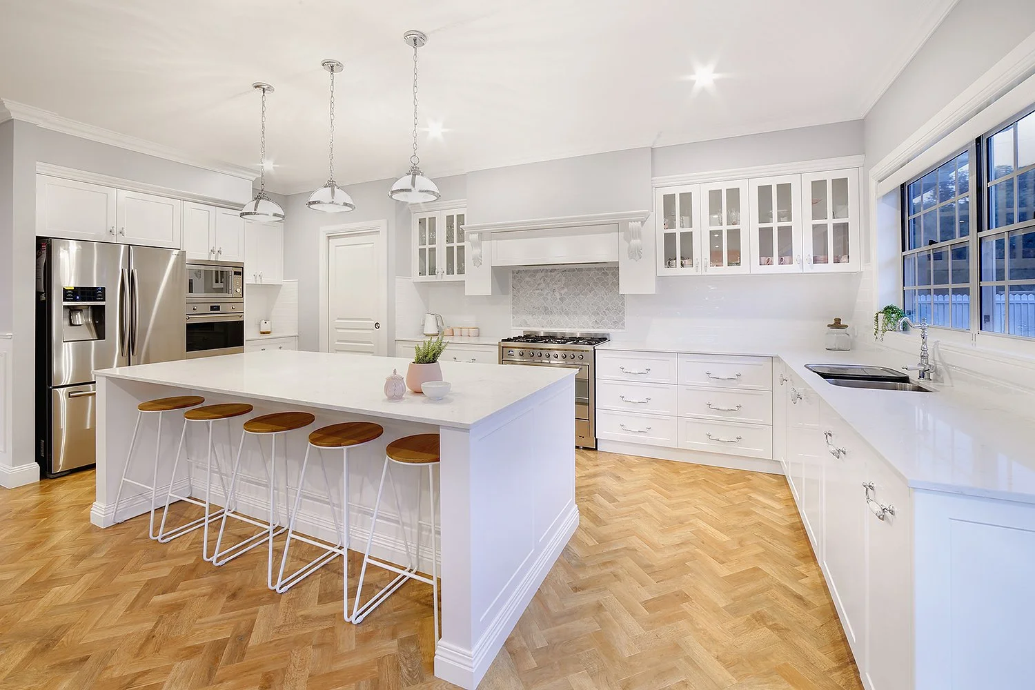 Bright white kitchen with stainless steel appliances, an island with four wooden stools, light hardwood floors, and a large window above the sink.