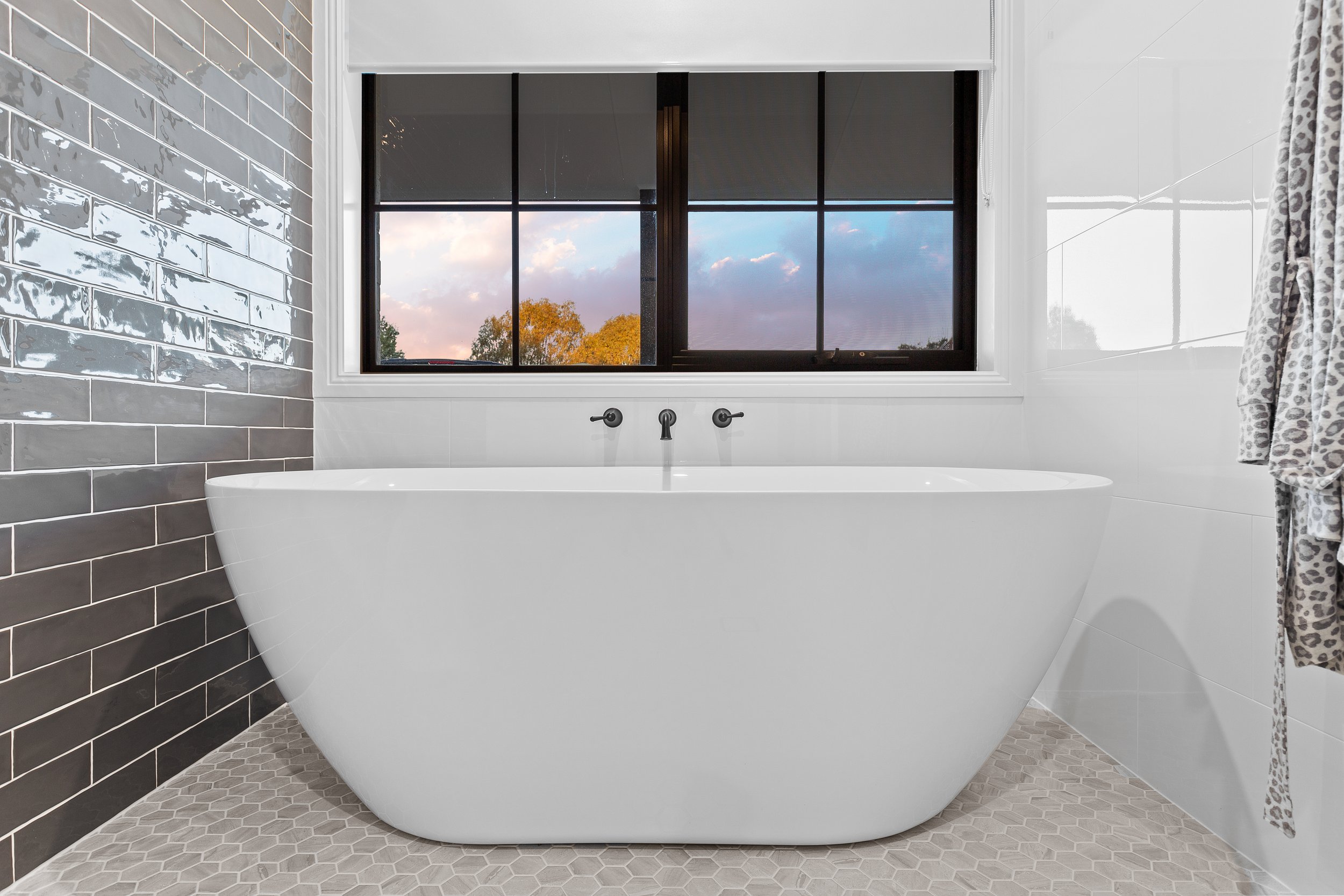 Bathroom with a white freestanding bathtub placed under a black-framed window showing a sunset sky and trees. The wall behind the bathtub has black subway tiles, and the floor has hexagonal beige tiles. A patterned towel hangs on the right side.