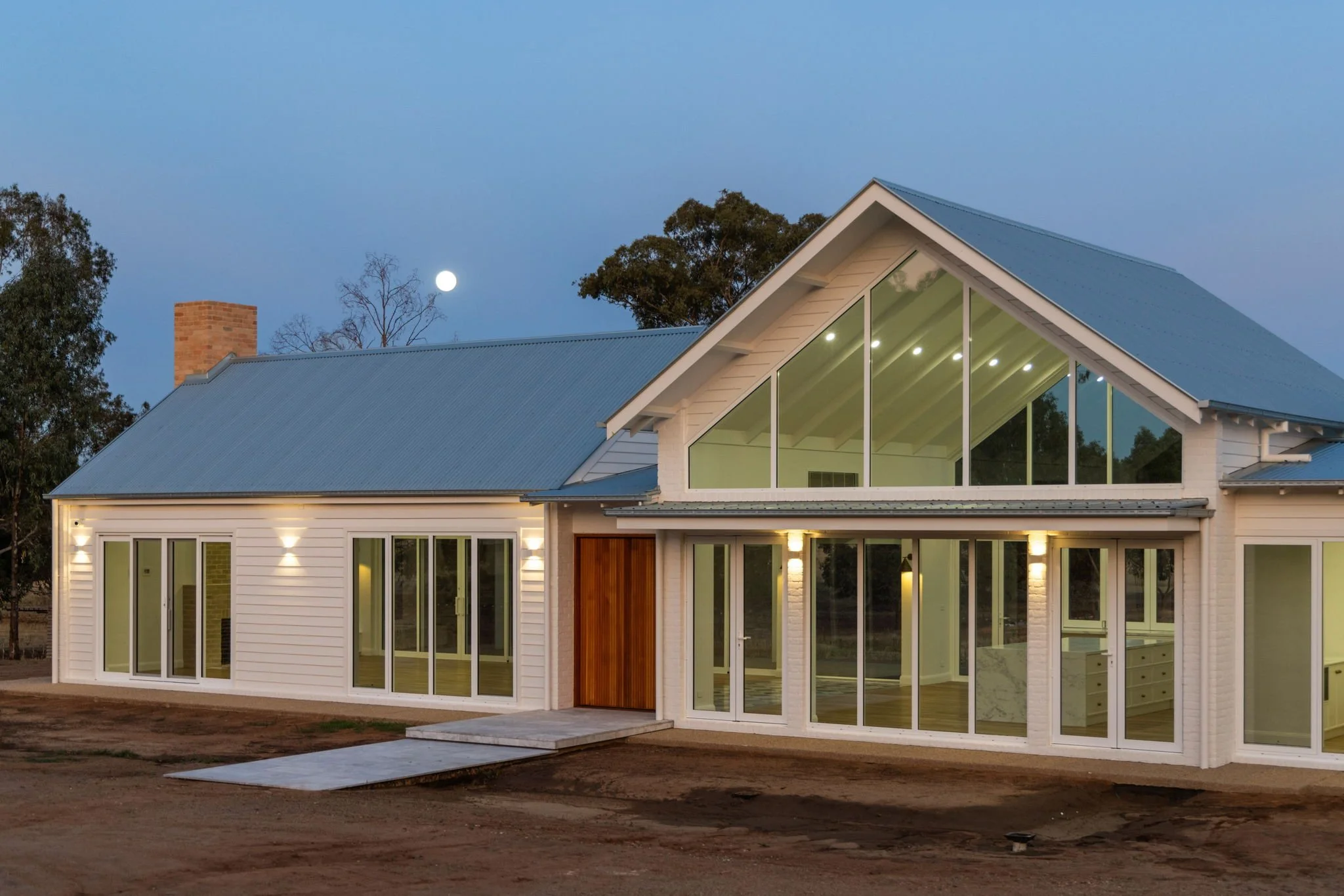 Modern house with large glass windows and white siding, front porch with a wooden door, surrounded by trees, nighttime with the moon visible in the sky.