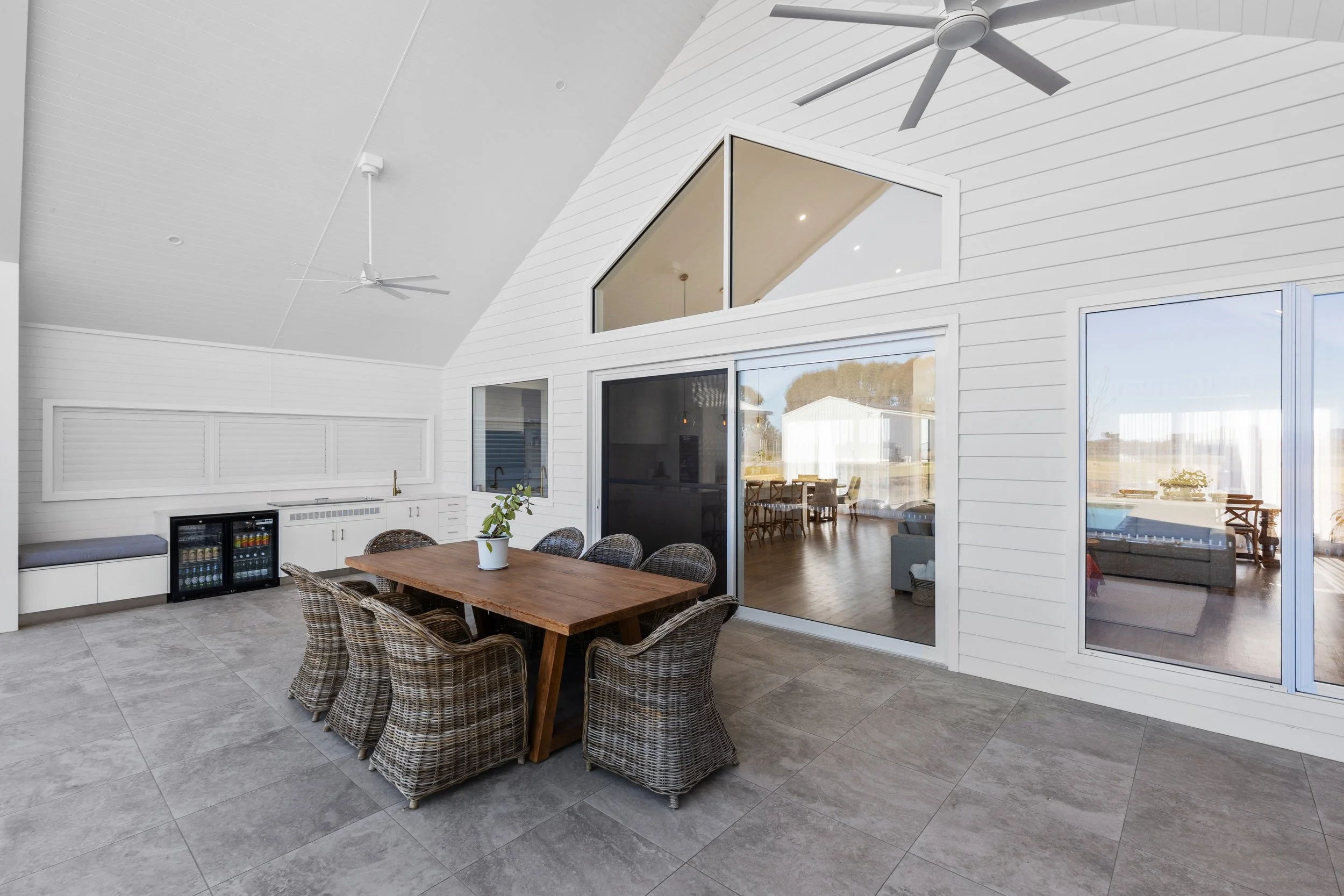 Covered outdoor patio with a wooden dining table, eight wicker chairs, a small plant in a white pot, built-in bench with cushion, mini fridge, white kitchen cabinets, ceiling fans, and large sliding glass doors leading to indoor living space.