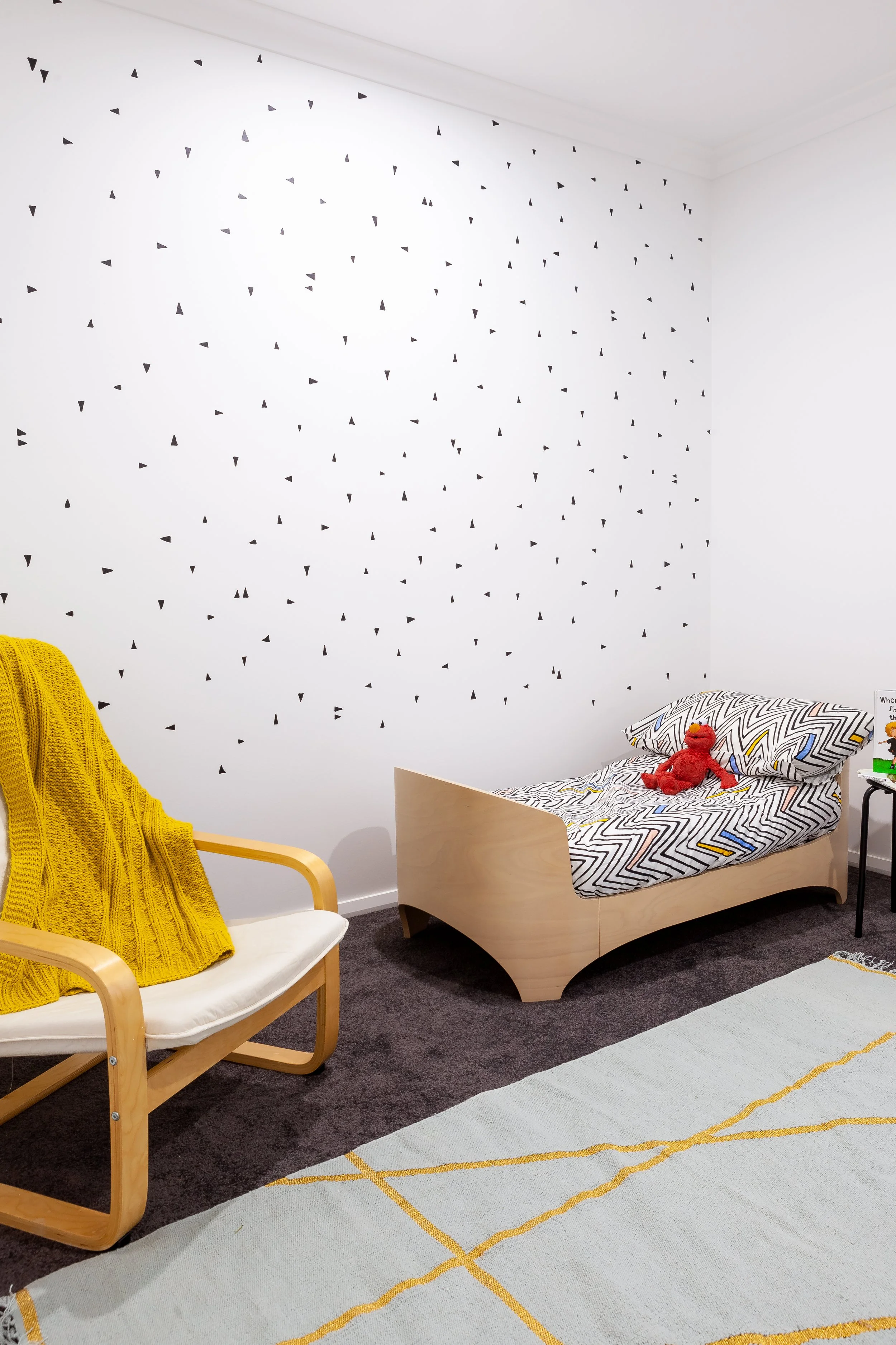 A child's bedroom with a small bed, a chair with a yellow blanket, a rug with yellow lines, and a stuffed red toy on the bed.