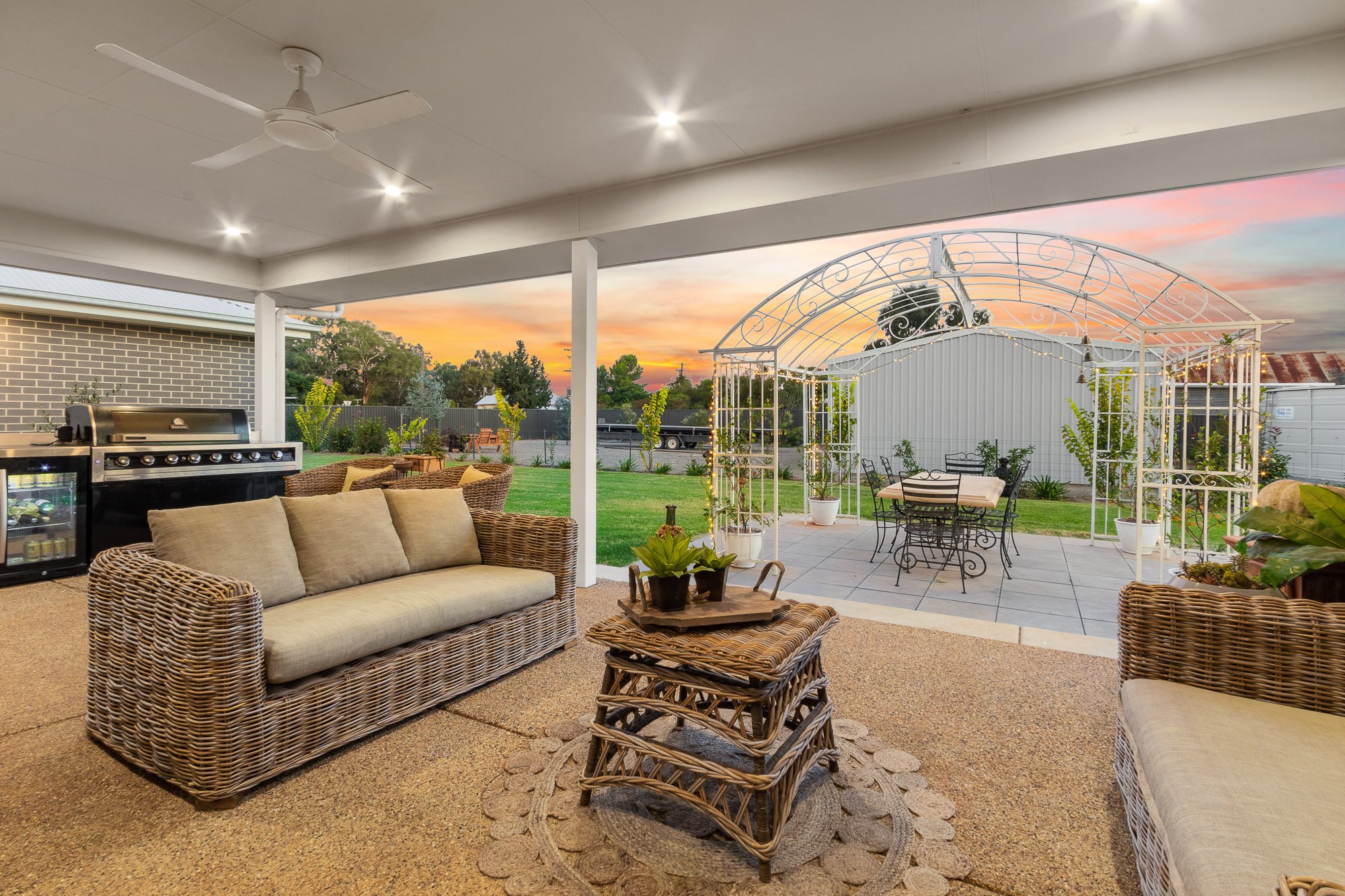 Covered patio area with wicker sofa and chairs, a small wicker table with plants, a grill, and an outdoor dining space under a white metal arbor with string lights, greenery, and a sunset sky in the background.