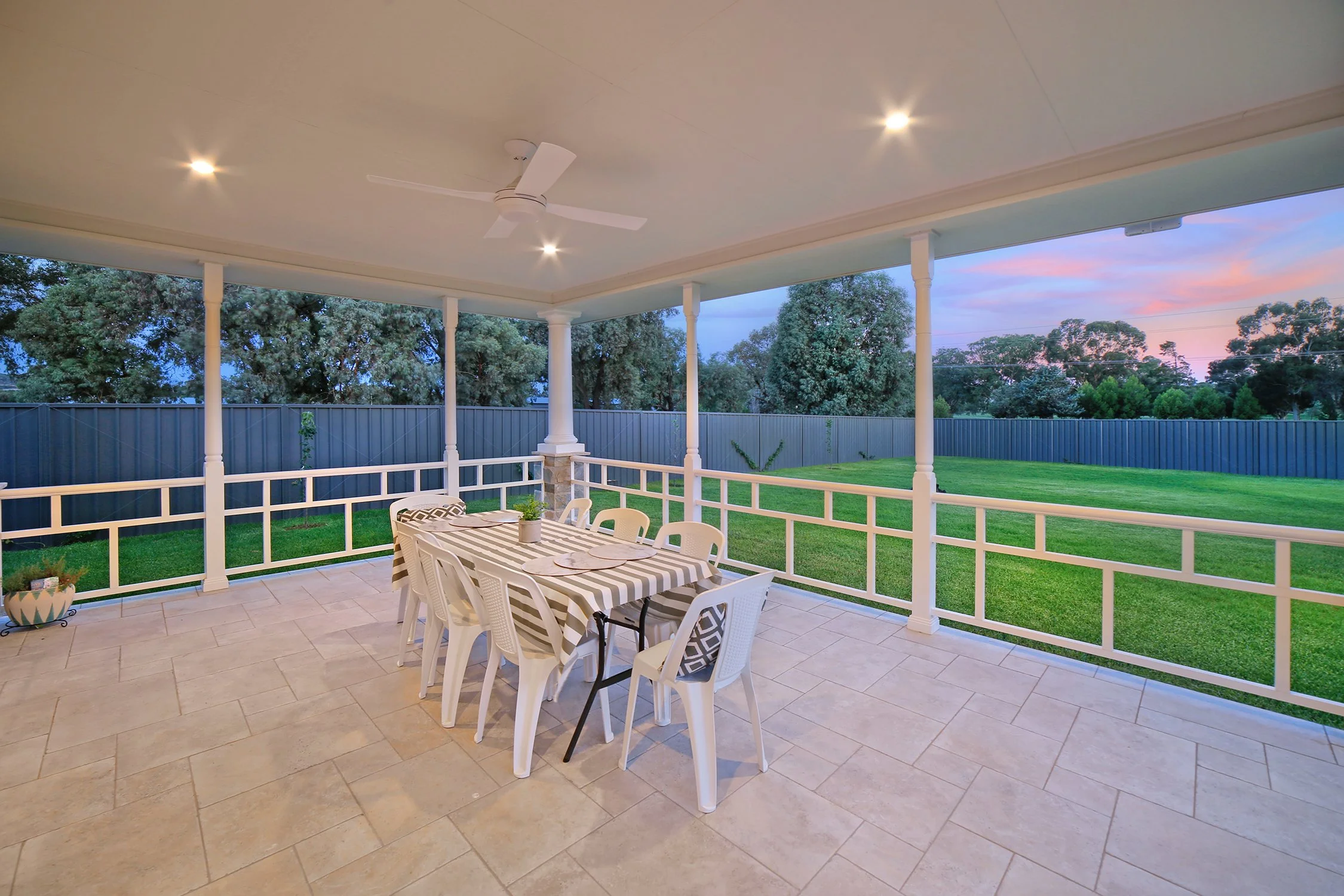 Covered porch with a dining table and six chairs, overlooking a grassy backyard with trees and a sunset sky.