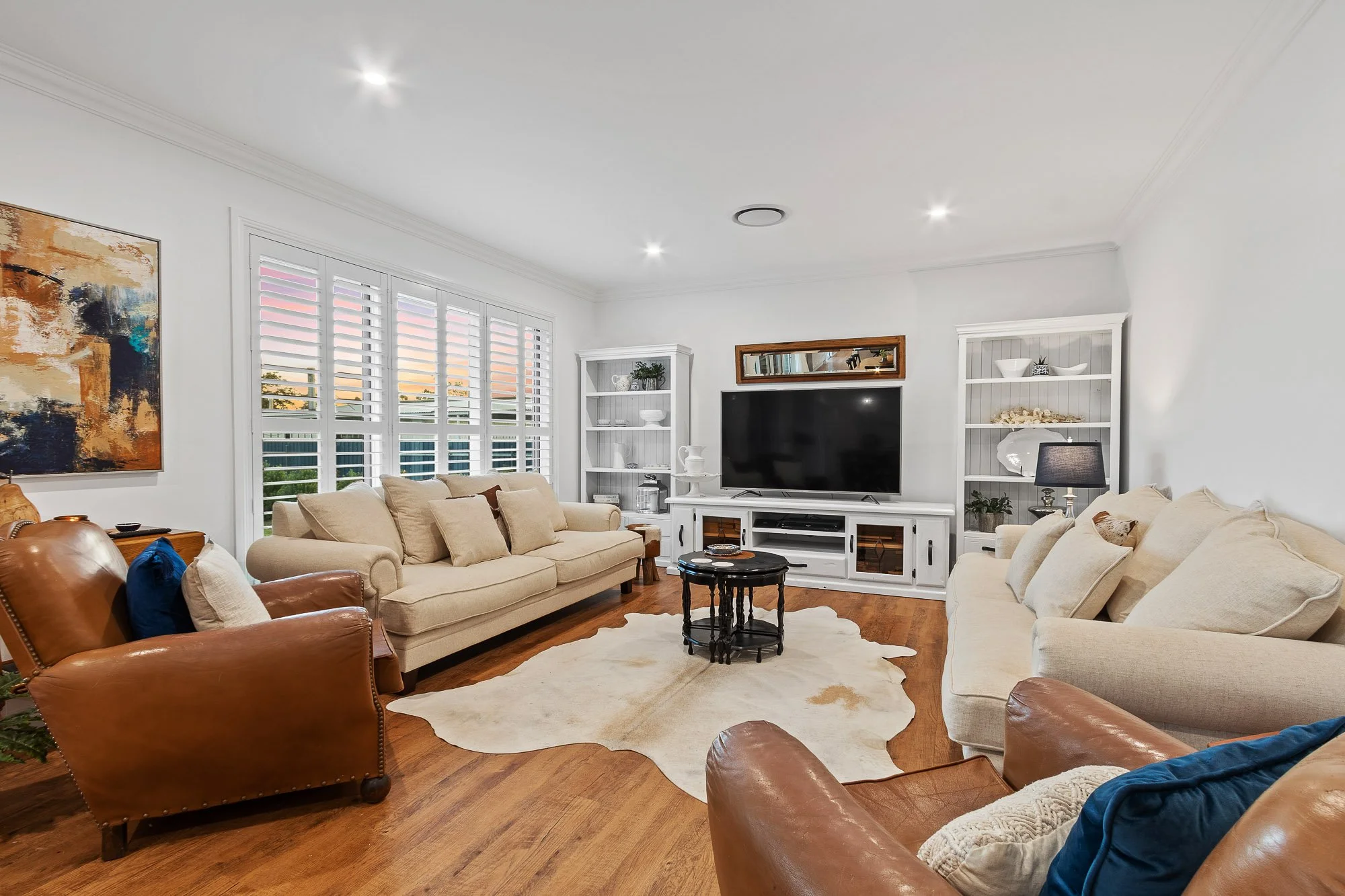 Living room with beige and brown sofas, wooden floors, white walls, a large window with white shutters, a white entertainment center, black TV, shelves with decorative items, and a cowhide rug.