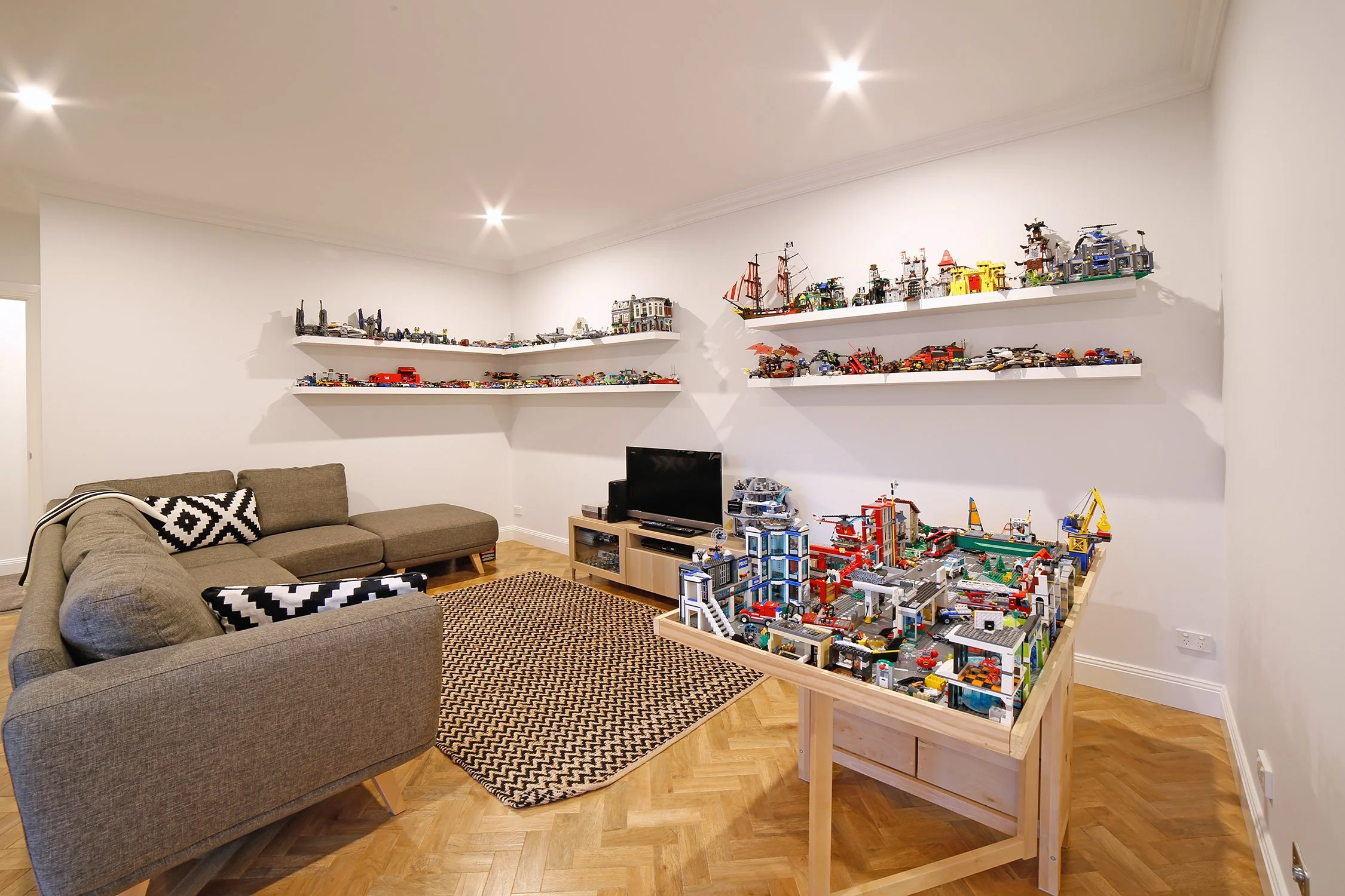Living room with beige sofa, black and white patterned pillows, a black and white rug, wooden herringbone floor, white walls with shelves of LEGO sets, a small TV on a wooden stand, and a large LEGO city display table.