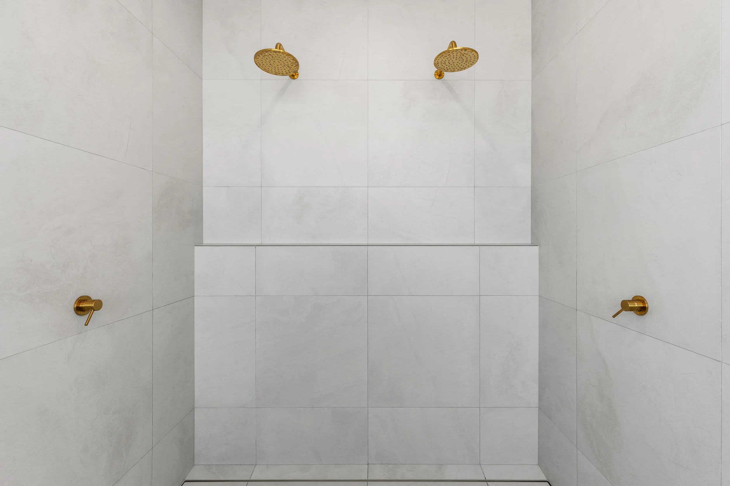 A modern shower with two overhead shower heads and two side control handles, all in a gold finish, on white tiled walls.