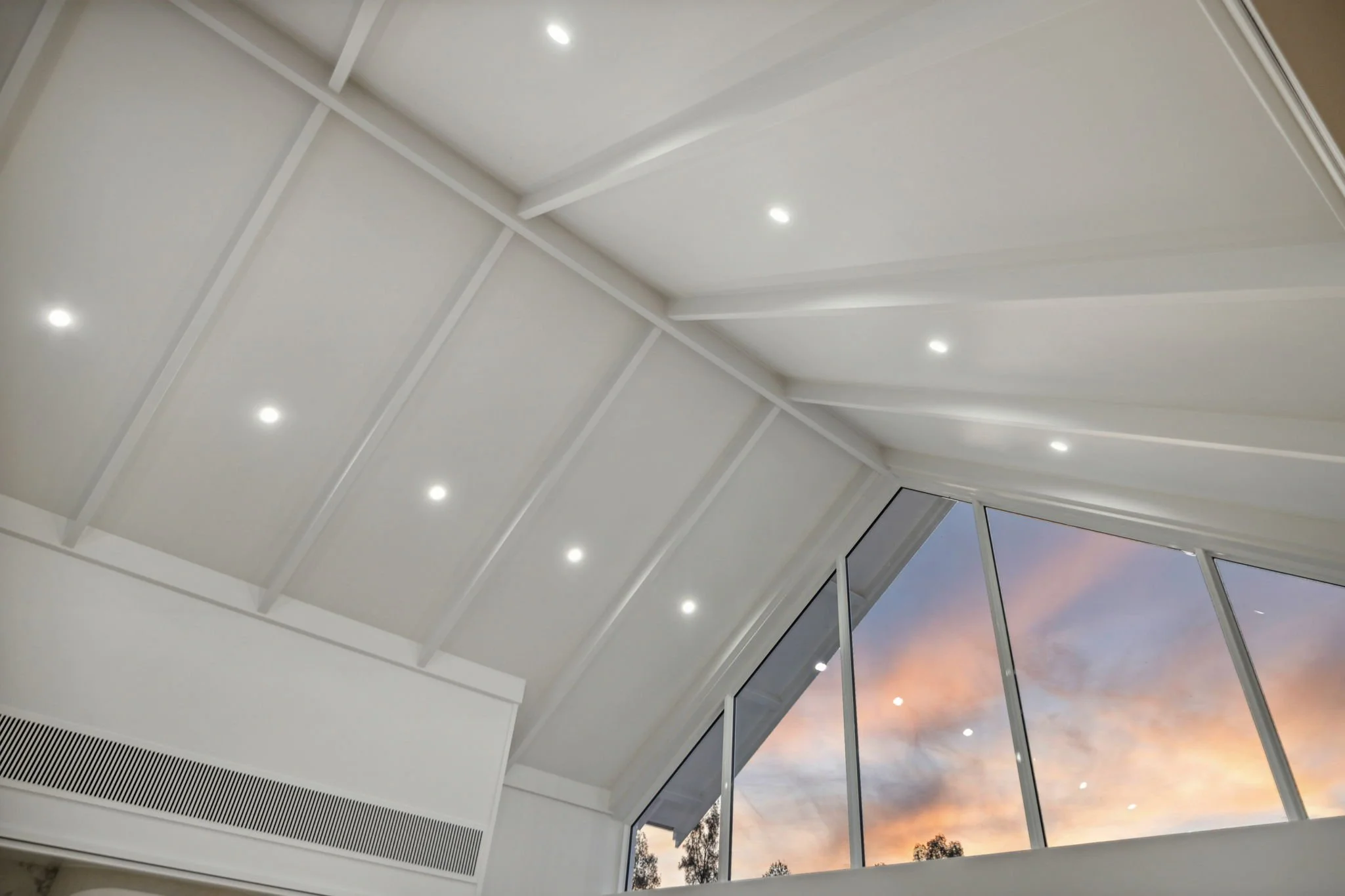Interior ceiling with white slats, ceiling lights, and a large triangular window showing a sunset sky with clouds and trees.