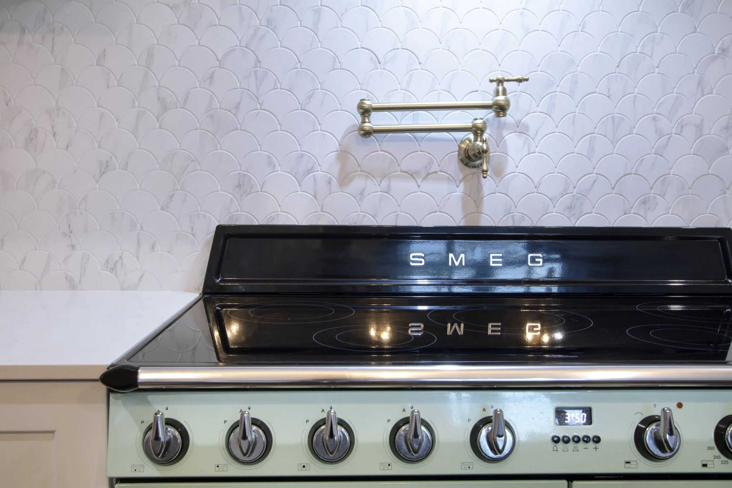 A vintage Smeg stovetop with four burners, control knobs, and a digital timer, set against a white tiled wall with a brass faucet above.